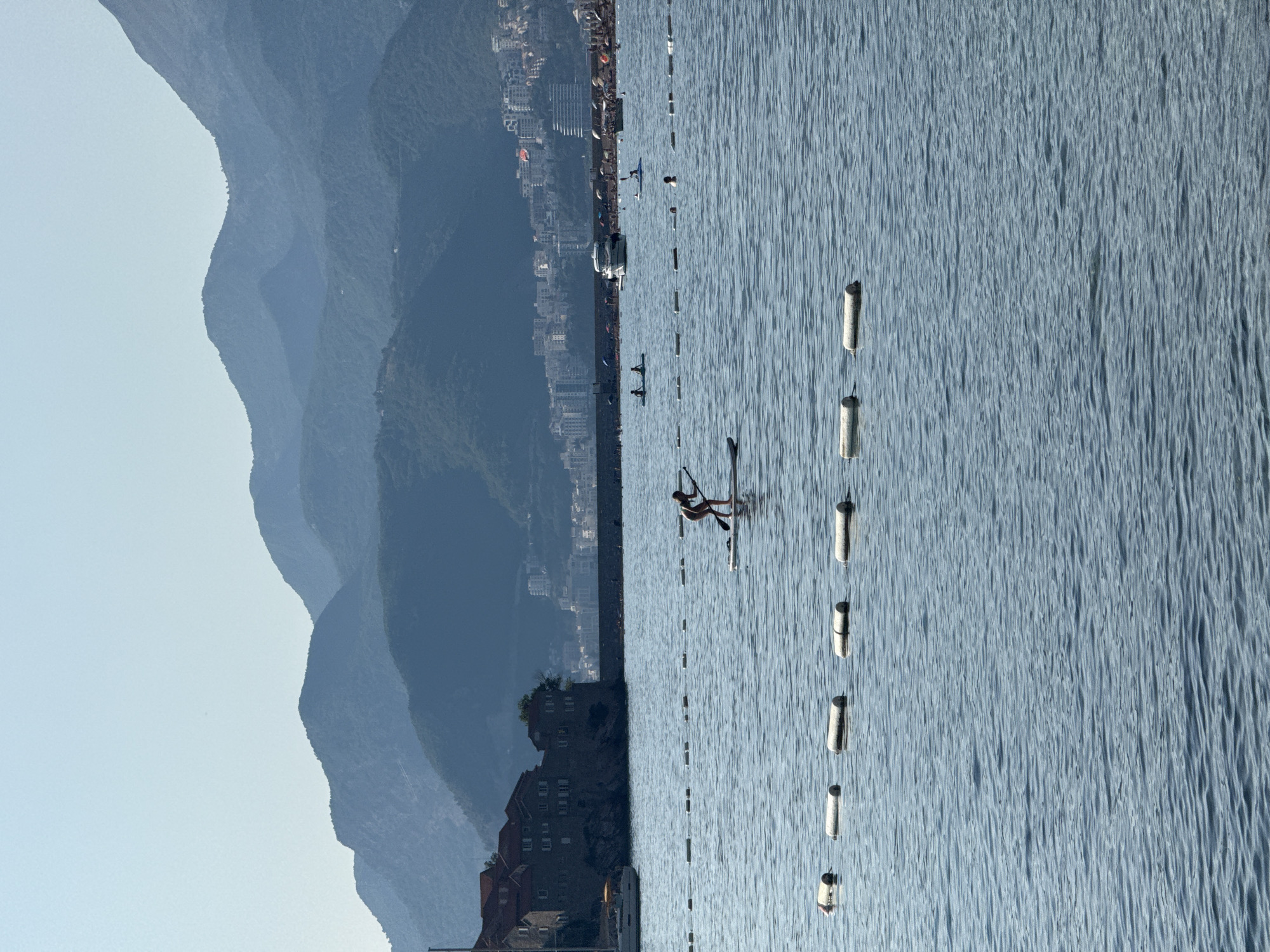 The image shows a body of water, likely the Adriatic Sea, with a person paddleboarding in the foreground. The paddleboarder is a solitary figure, standing and holding a paddle, facing away from the camera. In the mid-ground, a line of buoys floats on the water's surface. Beyond the buoys, a beach area is visible with numerous people, umbrellas, and a white boat anchored offshore. Further in the distance, a town or city with buildings lines the coast, backed by a series of layered mountains. The sky is clear and blue, indicating a sunny day. The architectural style of the buildings on the coast, particularly the dark-stone structures on a peninsula in the left mid-ground with a sailboat docked nearby, are consistent with Sveti Stefan, Montenegro. The layered mountains in the background are also characteristic of the mountainous terrain surrounding the Sveti Stefan region.