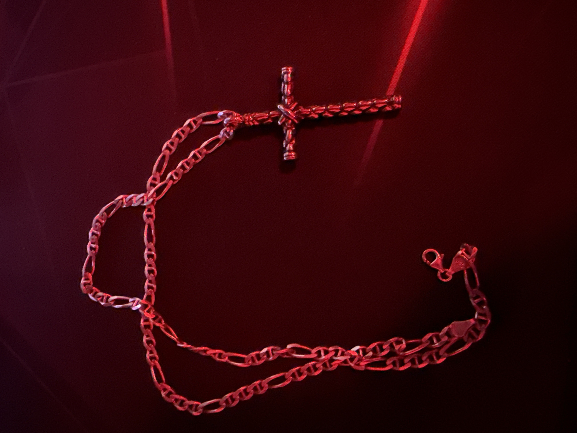 A cross necklace with a figaro chain lies on a dark red, glossy surface. The necklace is silver and has a polished finish. The cross is approximately 2 inches in length and features a textured pattern. The chain has a lobster clasp closure. The lighting in the image is red and casts highlights and shadows on the necklace and surface. There are no people or other objects visible in the image. The image was captured in Blagoevgrad, Bulgaria.