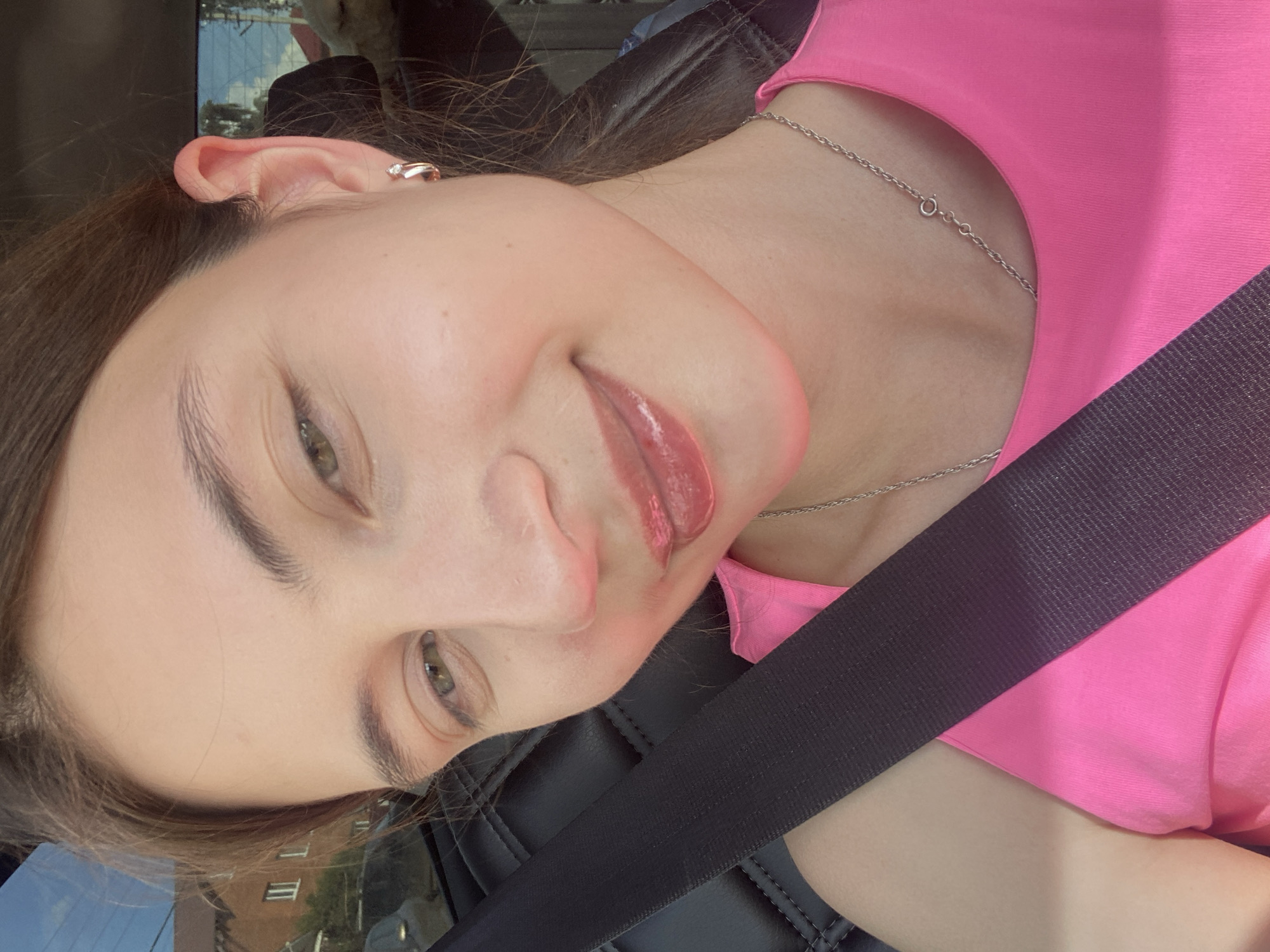A close-up, front-facing selfie shows a young woman with light skin, brown hair pulled back, and light green eyes, wearing a bright pink tank top and a silver necklace. She has on a subtle makeup, including lip gloss that gives her lips a shiny appearance. A small, silver earring with a tiny stone is visible in her right ear. A black seatbelt stretches across her chest, indicating she is inside a vehicle. The background is blurred, but suggests an outdoor setting with hints of buildings and trees, possibly a residential area in or near a city like Kamkoye, Russia. The lighting is bright, suggesting it is daytime.