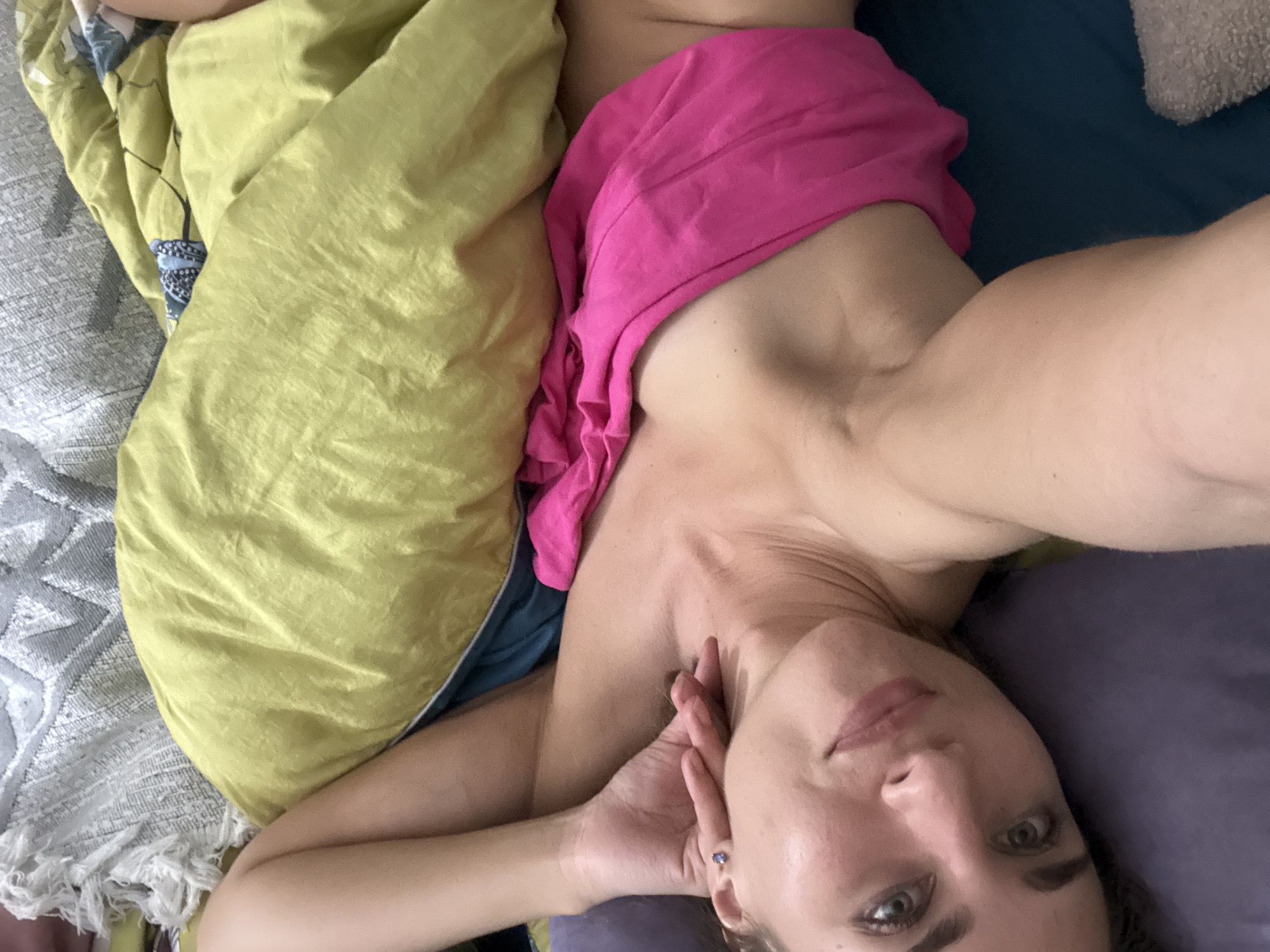 A person is lying in bed, covered partially by a yellow blanket and a pink garment. Their face is visible, with eyes open and looking towards the camera. They are wearing a small blue earring. The scene appears to be indoors, likely a bedroom, with pillows and bedding visible. The lighting suggests it could be daytime, but there are no strong indicators of time of day or weather. The person's expression is neutral. There is no visible text.