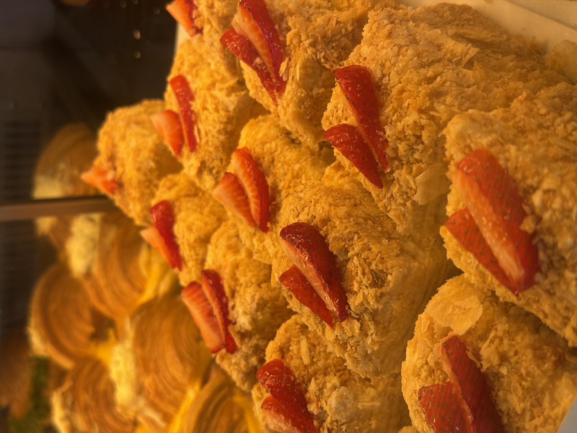 The image displays a close-up view of freshly baked pastries arranged in a display case, likely in a bakery or cafe. The primary subjects are multiple individual portions of what appear to be Napoleon cakes (Mille-feuille), characterized by their flaky, golden-brown crust and crumbly texture. Each pastry is rectangular or square-shaped and generously topped with several bright red slices of fresh strawberries, indicating a fruit-flavored or garnished dessert.
These strawberry-topped pastries are positioned in the foreground and mid-ground, appearing stacked or laid out in rows, drawing the viewer's attention to their intricate textures and vibrant fruit. The lighting is warm, enhancing the golden hues of the pastry and the rich red of the strawberries.
In the blurred background, another type of pastry is visible. These appear to be spiral-shaped baked goods, possibly cinnamon rolls or similar coiled pastries, suggesting a variety of offerings in the establishment. The depth of field is shallow, keeping the focus sharply on the strawberry cakes while the background elements are softly out of focus. No people or visible text are discernible in the image. The scene suggests a retail display, showcasing tempting desserts under artificial light.