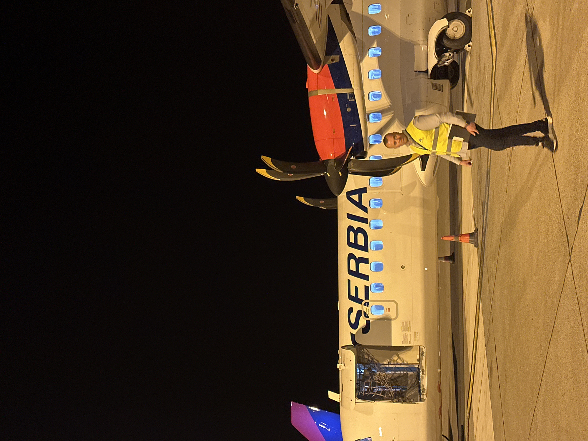 A worker in a yellow safety vest stands next to an Air Serbia aircraft on a tarmac at night. The word "SERBIA" is vertically aligned on the plane's fuselage. The aircraft's turboprop engine is visible. An orange traffic cone is near the aircraft. The location is likely Podgorica, Montenegro, based on the airline and typical airport operations. The scene is illuminated by artificial lights.