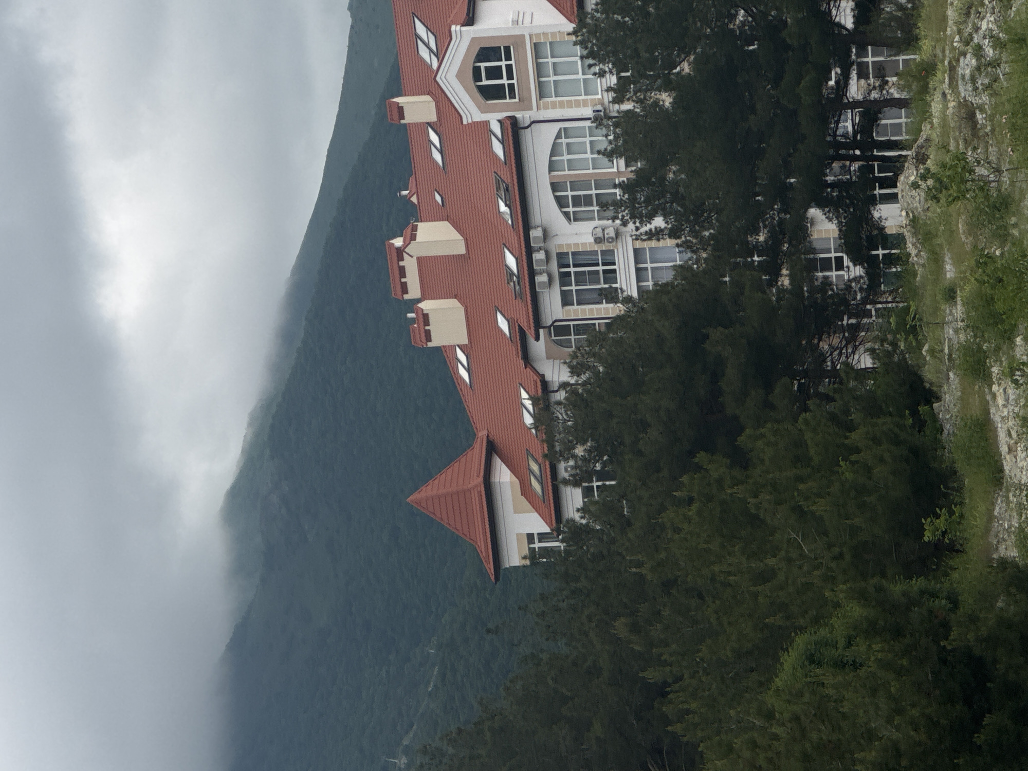 The image presents a partial view of the city of Gelendzhik, Russia, framed by a mountainous landscape. A portion of a multi-story building with a distinctive red tiled roof occupies a significant portion of the frame. The building's architectural style includes a combination of traditional and modern elements, as evidenced by the contrasting windows and facade. Dense foliage of trees partially obscures the building, suggesting a wooded or landscaped setting. In the background, a mountain range is partially enveloped in mist or low-hanging clouds, adding a sense of depth and atmosphere to the scene. The lighting appears soft, suggesting an overcast or early morning condition.
