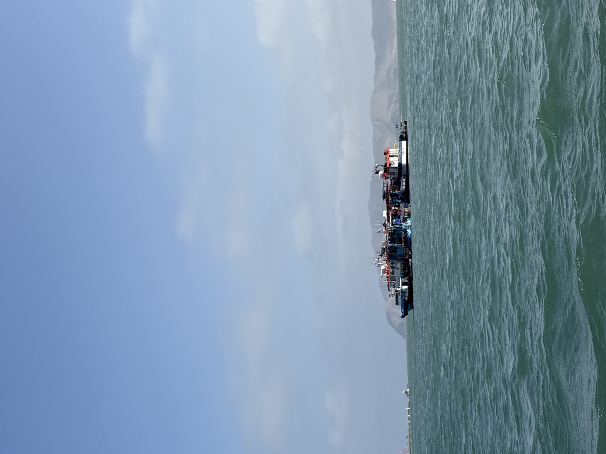 The image shows a series of boats docked in the water, presumably in the city of La Punta, Peru, given the prompt's location information. The water is choppy. There is a mountain range in the distance, and the sky is mostly clear.