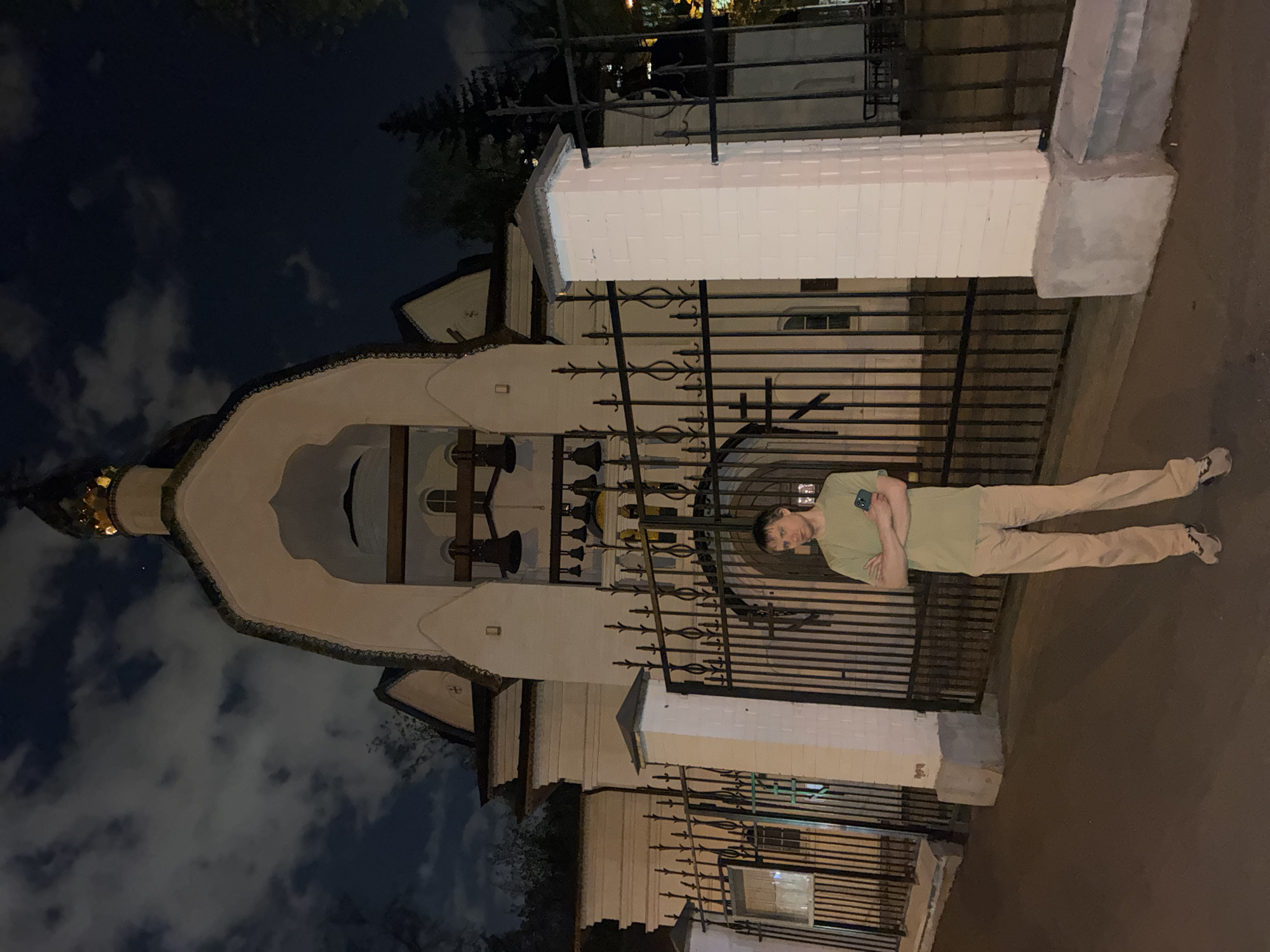 The image shows a man standing in front of the Danilov Monastery in Moscow, Russia, at night. The monastery is lit up, and the sky is visible with clouds. The man is wearing a green shirt and beige pants. He is standing with his arms crossed. A black fence surrounds the monastery.