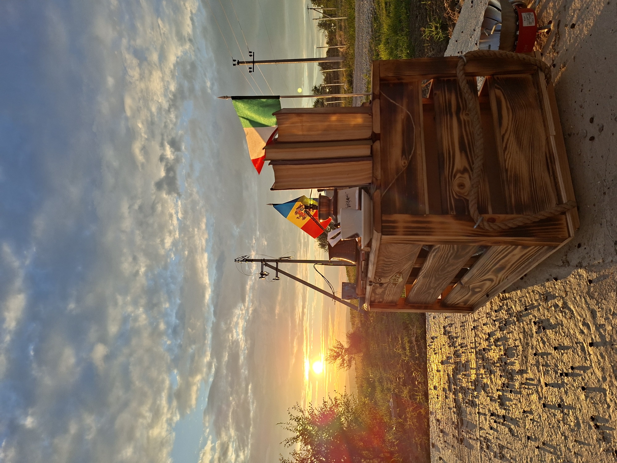 A vertical, eye-level, medium shot captures a rustic wooden crate on a concrete surface, set against a backdrop of a sunset in Sociteni, Moldova. The sun, a bright orange orb, is partially obscured by clouds and casts a warm glow across the landscape. The sky is filled with dramatic, wispy clouds, ranging in color from pale blue to golden yellow. In the foreground, the wooden crate is adorned with two small flags, one of Italy and one of Moldova, and several books. A white teacup and saucer sit on top of the crate. To the left, a bushy tree with illuminated foliage stands tall. In the distance, power lines and poles stretch across the horizon, with a hint of rural buildings visible. The concrete surface in the foreground is textured and scattered with dark particles, casting long shadows in the warm sunlight. The overall scene evokes a sense of peacefulness and natural beauty.