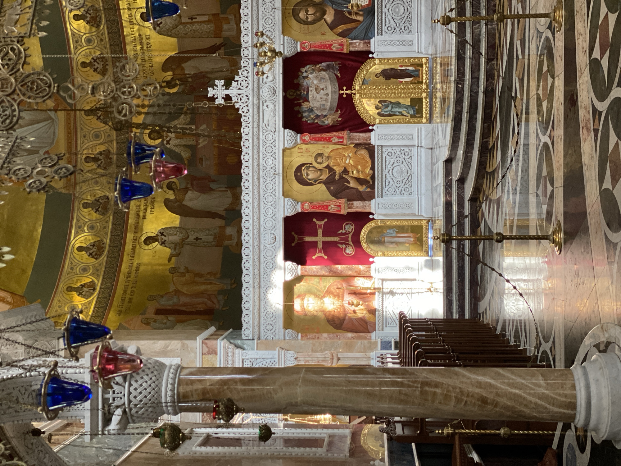 The image captures the interior of a Russian Orthodox church, likely located in Yekaterinburg, Russia. The focus is on the iconostasis, a wall of icons that separates the sanctuary from the nave. The iconostasis is intricately decorated with gold and white carvings, and features religious icons depicting various saints and biblical scenes. A chandelier with colored glass lamps hangs in the foreground. The floor is patterned with geometric designs in dark and light stone. Pews can be seen to the right, along with gold-colored stands with chains hanging from them. The walls are adorned with religious murals and script. A marble column with a white capital is visible in the foreground.
