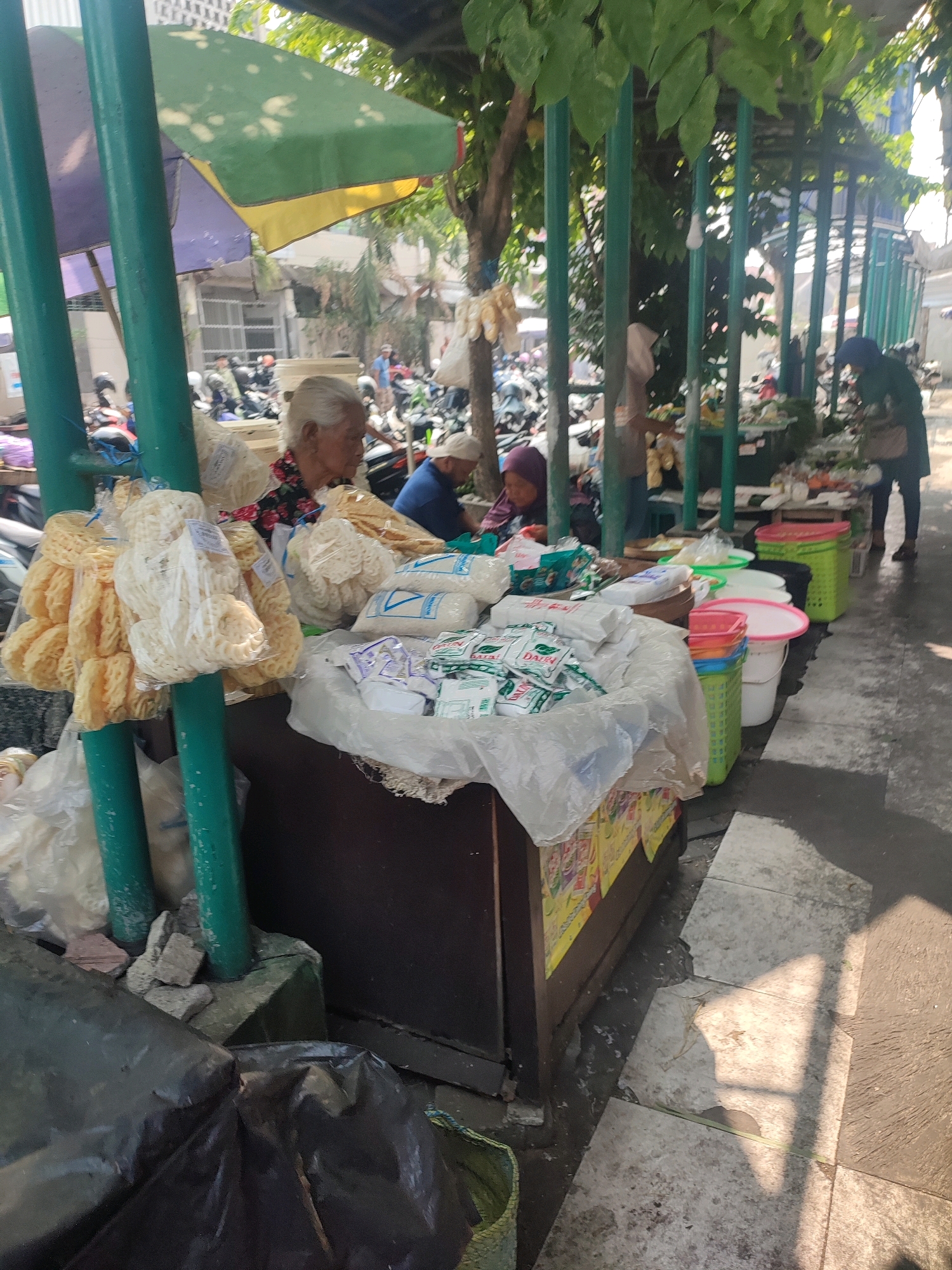 A street market in Yogyakarta, Indonesia, features vendors and customers under makeshift awnings and stalls. An elderly woman, wearing a floral shirt and a headscarf, sits behind a display of packaged snacks and food items. In front of her, several plastic bags contain assorted fried crackers or chips. Behind her, a man in a blue shirt and a white cap is visible. The market setting includes motorcycles parked in the background, and various plastic containers, bags, and other goods are laid out on tables and the ground. Sunlight casts shadows on the pavement, indicating an outdoor setting.