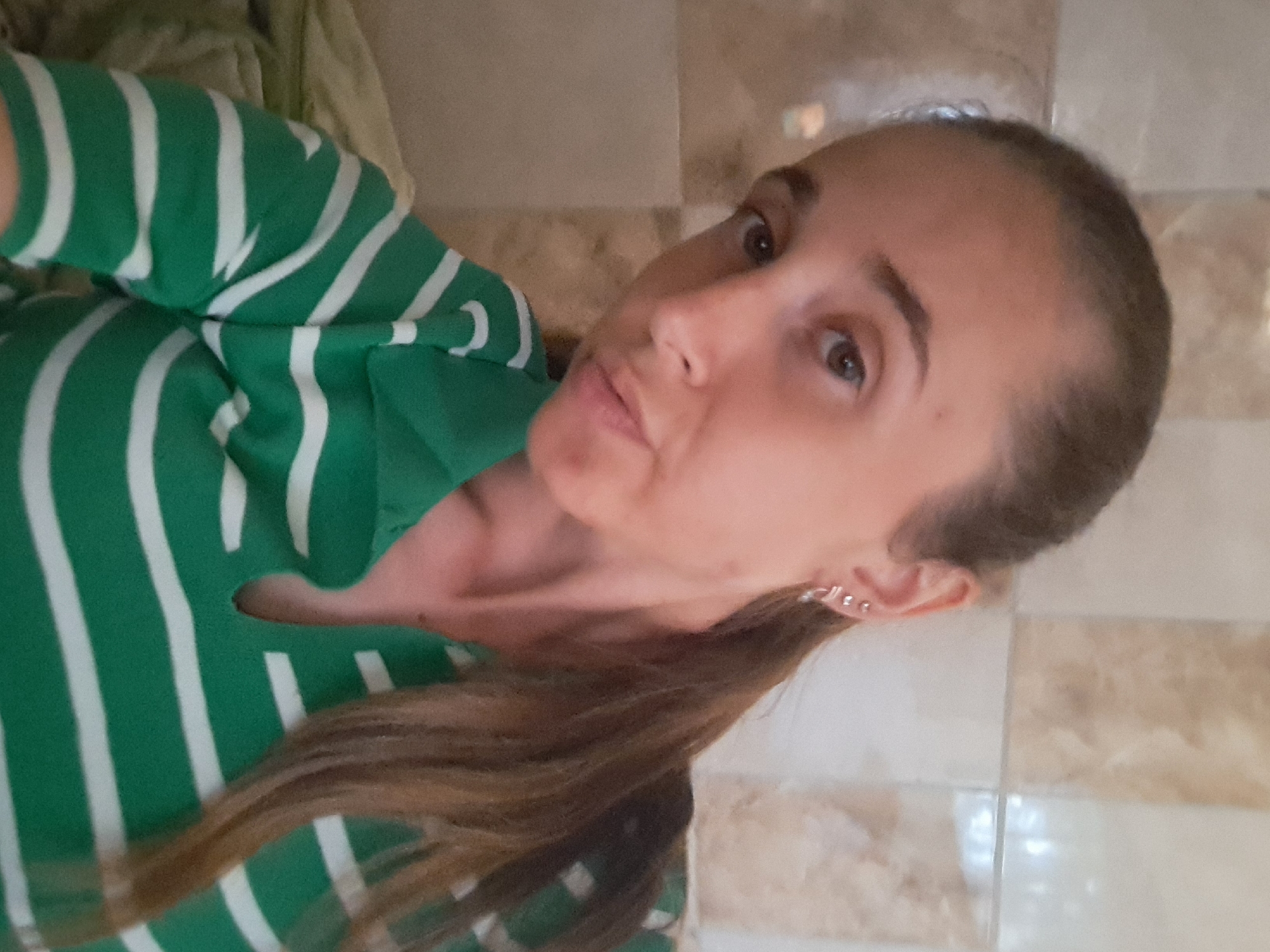 A young woman with long brown hair pulled back in a ponytail is taking a selfie. She is wearing a green and white striped polo shirt. She has earrings in her ears. The background appears to be a tiled wall. The image is a close-up, showing her face and upper body. There is no discernible location within the city of Vypasne, Ukraine.