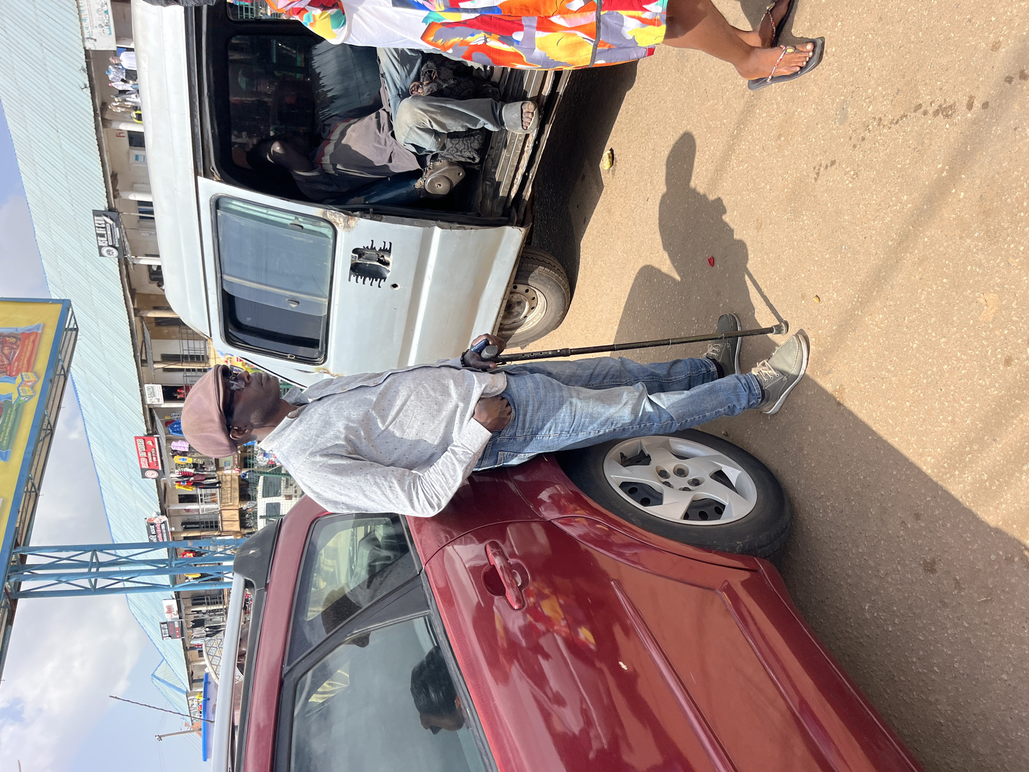 In an outdoor urban setting under a bright sky, a man stands next to a dark red car, leaning on a cane. He wears a light-colored, long-sleeved collared shirt, blue jeans, a flat cap, and sunglasses. To his right, a silver minivan is parked, its sliding door open. Inside the minivan, a person is visible sitting down. Further to the right, the lower half of another person wearing a colorful dress and flip-flops is visible. The ground is paved, and in the background, buildings with blue roofs and shopfronts line a street. There are overhead signs and structures, suggesting a market or commercial area. The scene is lit by sunlight, casting shadows on the ground.
