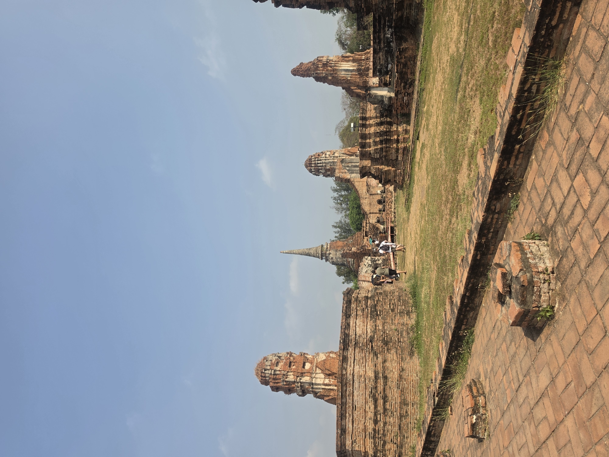 The image depicts an ancient temple complex in Phra Nakhon Si Ayutthaya City Municipality, Thailand. The scene features a mix of red brick structures, including ruined chedis and walls, against a backdrop of a clear blue sky with light clouds. Some of the structures are in disrepair. A grassy field extends through the complex. Several tourists are visible in the middle ground, some walking and others standing near the ruins. The foreground includes a brick-paved area and a low brick wall.