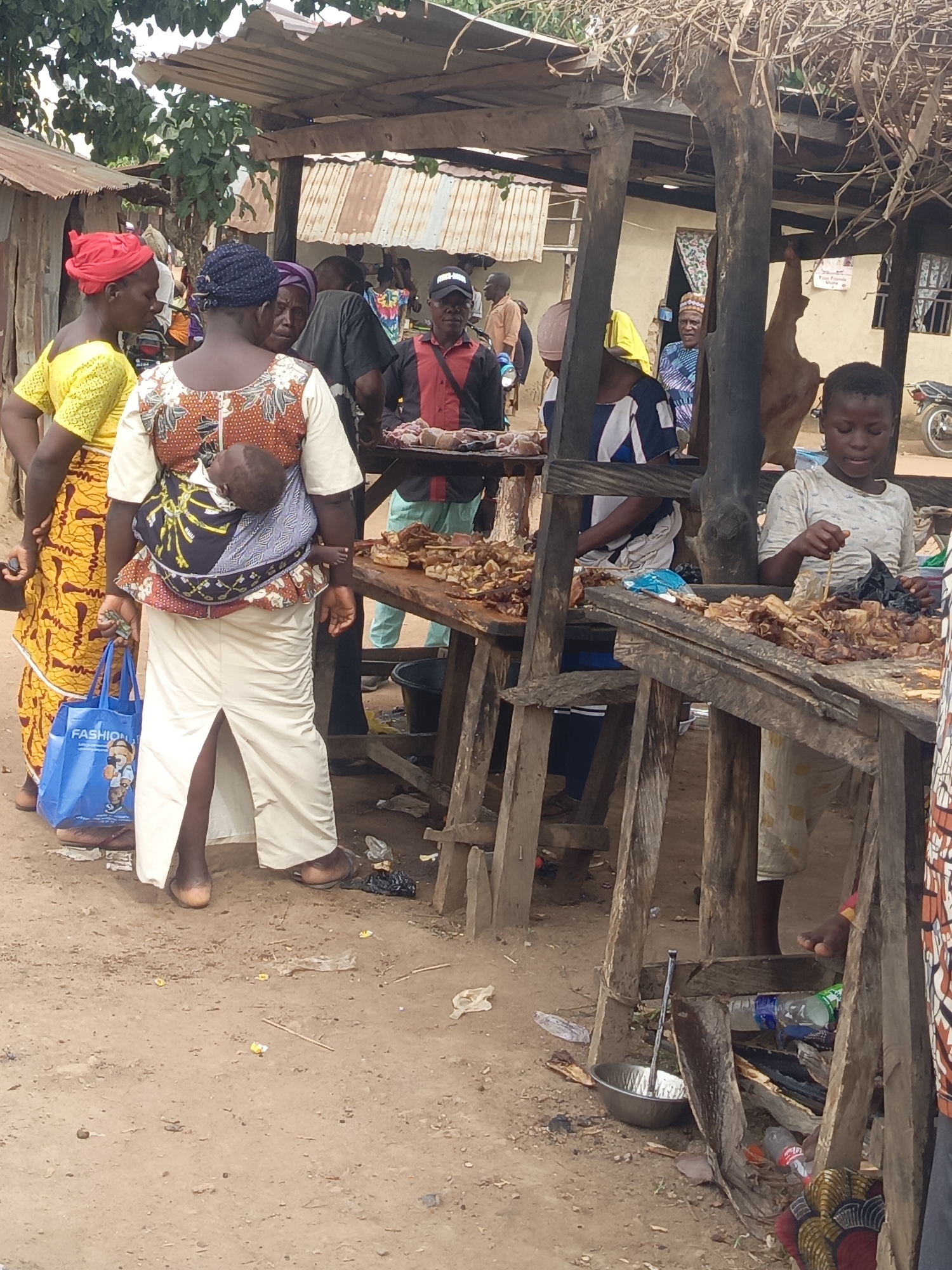 This daytime image captures a vibrant outdoor market scene in Andaha, Nigeria. The setting is characterized by rustic wooden market stalls with corrugated metal or thatched roofs, and a dirt ground scattered with debris.
Numerous people are present, indicating a busy commercial environment. In the foreground left, a woman in a bright yellow dress and a red headwrap holds a blue shopping bag with "FASHION" written on it. Next to her, another woman is seen from behind, carrying a baby securely in a colorful patterned cloth carrier on her back. She wears a patterned top and a long, light-colored skirt.
The market stalls are laden with what appears to be roasted or grilled meat, suggesting food sales. To the right, a young child, likely a boy, stands behind a stall, engaged in sorting or preparing the food with a stick. Other adults are visible throughout the market, some observing the goods, others interacting.
Notable background details include simple buildings constructed from corrugated metal, trees, and a glimpse of motorcycles. A dark, elongated object resembling an animal carcass or hide hangs from a wooden post in the mid-ground, further emphasizing the nature of the market. The bright lighting suggests it is daytime. The overall scene depicts a functional and bustling community marketplace.
