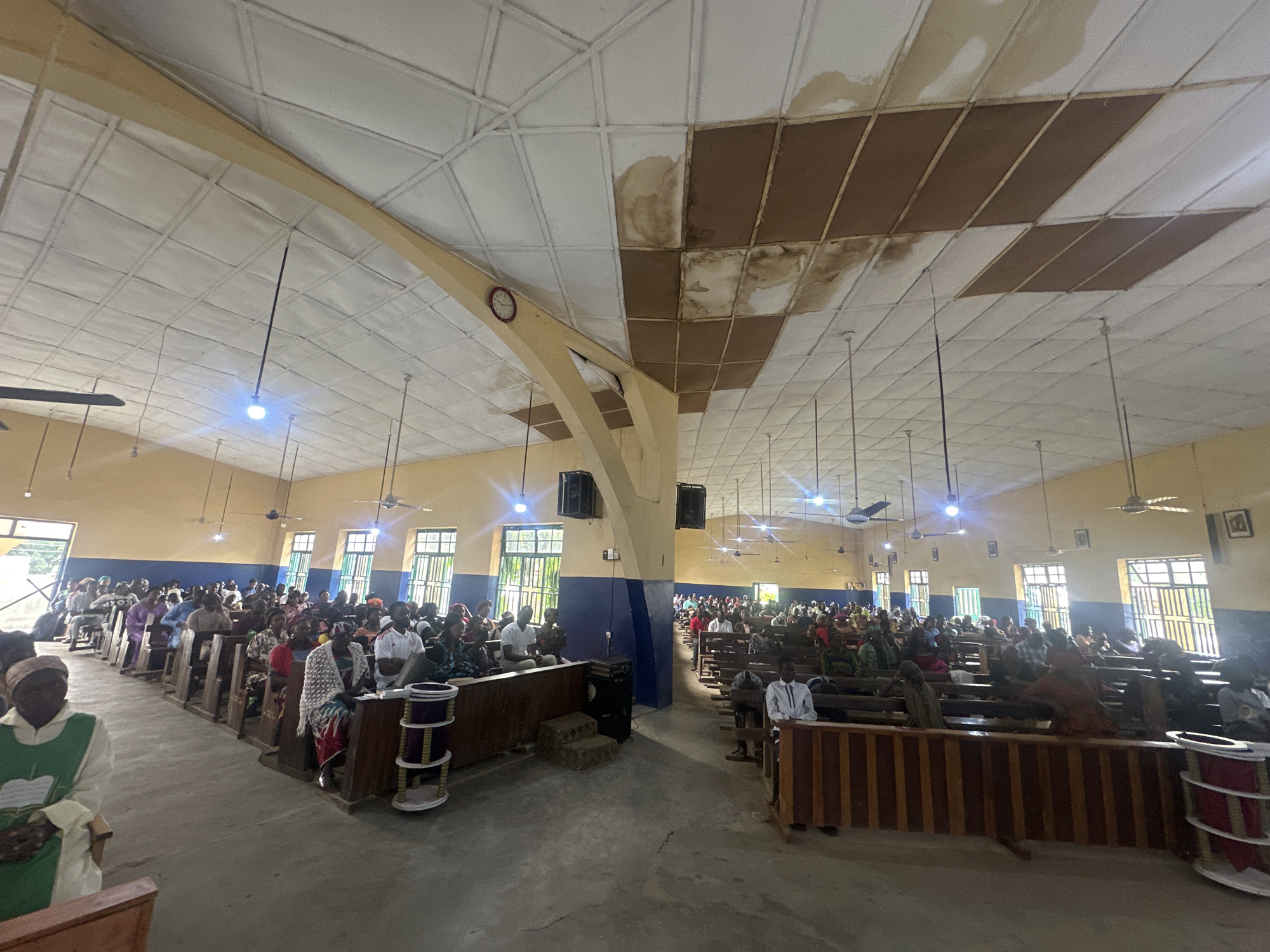 The image depicts the interior of a church in Akwanga, Nigeria, filled with a congregation seated on wooden pews. The church has a high, tiled ceiling with some water damage and discolored panels. Overhead fans and lights are visible, as are wall-mounted speakers and a clock on a prominent yellow pillar. The walls are painted in contrasting colors of yellow and blue. Sunlight streams in through barred windows. People of various ages are dressed in diverse attire, some wearing traditional clothing. A woman in a green vestment is in the foreground on the left. The overall atmosphere appears to be one of a religious gathering, possibly a service or meeting.