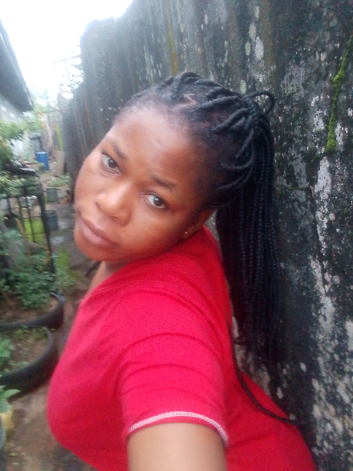 A medium shot captures a young Black woman with braided hair, wearing a red top, in an outdoor setting. She is positioned in the foreground, her head tilted to her left, looking directly at the camera. Her skin tone is brown, and her eyes are brown. Her hair is styled in long, dark braids that fall over her right shoulder. The red top features a white trim around the neckline.
The background is blurred and appears to be a somewhat unkempt outdoor area. To the left, there are hints of greenery, possibly plants in pots or a garden, and a corrugated metal structure, likely part of a building or fence. To the right, a textured, grey wall, possibly concrete or stone, is visible, partially covered with green moss and white splotches, suggesting it has been exposed to the elements. The overall lighting is soft and diffused, indicating an overcast or cloudy day. The environment suggests a residential area, possibly in Yenagoa, Nigeria, given the visual cues of the architecture and vegetation.