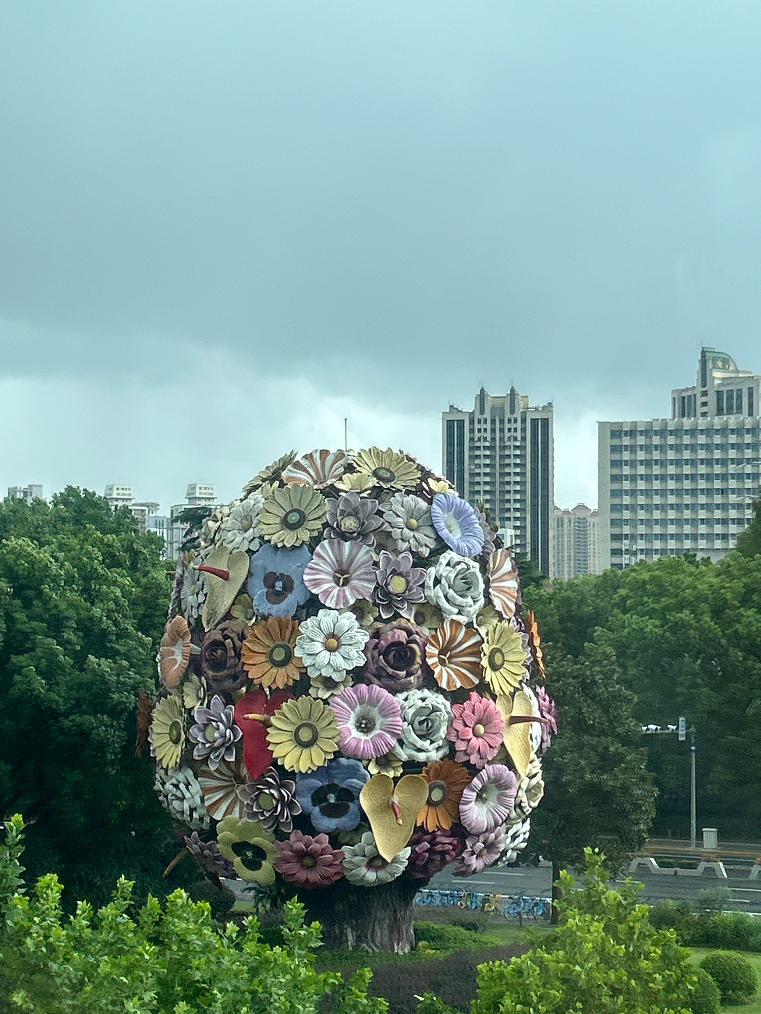 A large, egg-shaped sculpture covered in a mosaic of colorful, artificial flowers stands in the foreground, partially obscuring the view of a city skyline. The sculpture, appearing to be made of metal or ceramic flowers, displays a variety of flower types and colors, including shades of yellow, orange, pink, purple, blue, and white. Some flowers are layered, creating a textured and voluminous effect. A dark, tree-trunk-like base supports the floral egg.
Behind the sculpture, a dense canopy of green trees stretches across the middle ground, softening the urban landscape. In the background, several tall buildings rise into a overcast, pale blue sky. The architecture of the buildings suggests a modern city with a mix of styles, some featuring grid-like window patterns and others with more ornate tops.
The overall environment appears to be a public park or urban green space, given the presence of trees and what looks like a paved pathway or road with some bicycles parked in the distance. The weather seems to be cloudy or overcast, contributing to the diffused light. The image appears to be taken from a slightly elevated viewpoint, looking down and across the scene. This scene is located in the Changning District of China.