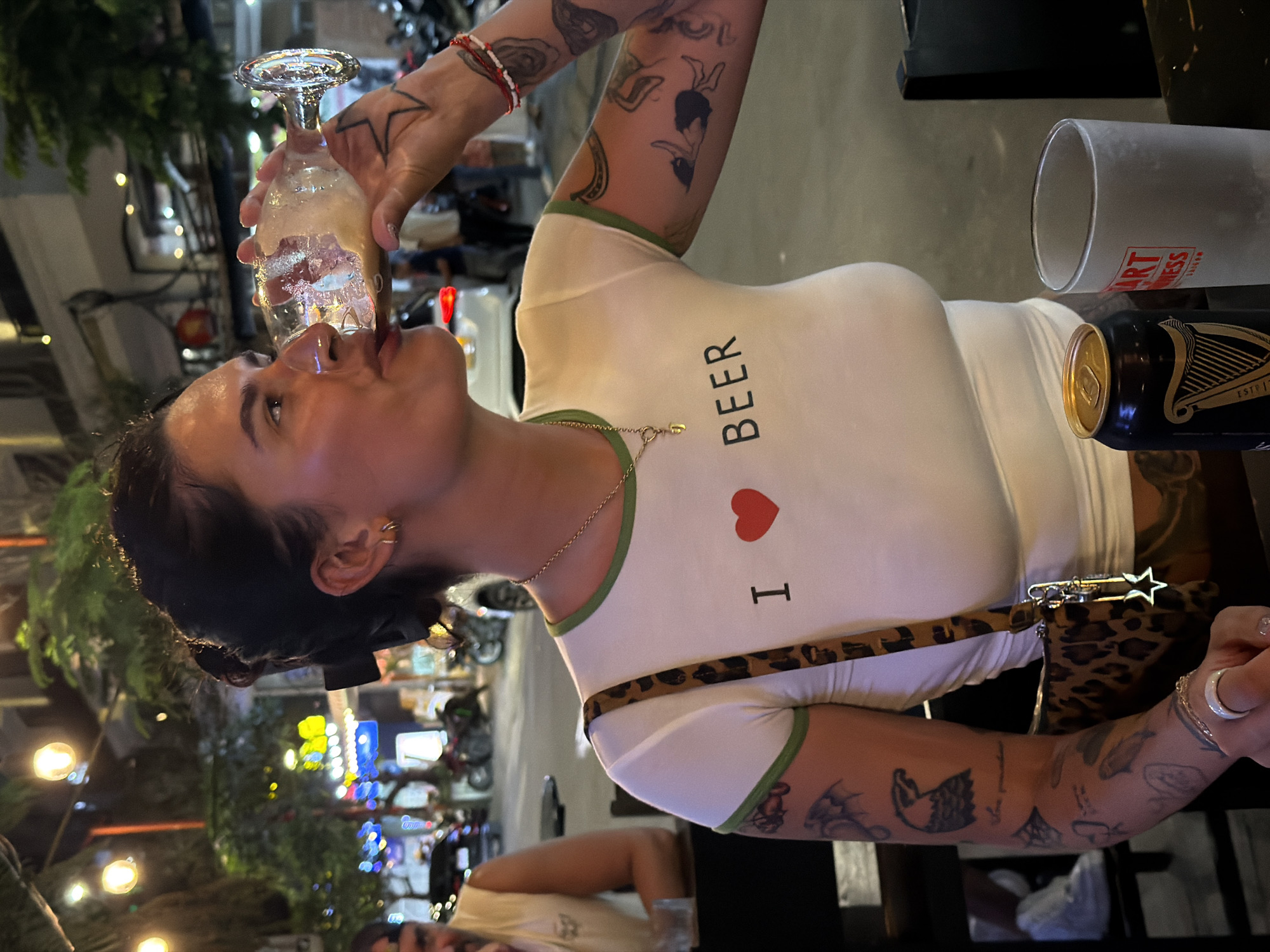 The image shows a woman drinking from a glass on a city street in Đà Nẵng, Vietnam. She has dark hair pulled back and is wearing a white t-shirt with green trim, a gold necklace, and a leopard-print purse. The shirt has the words "I <3 BEER" printed on the front. She has tattoos on her arms and is holding a glass up to her mouth. Next to her is a can of Guinness and an empty glass. The background shows a busy street scene with motorcycles and buildings lit up at night.