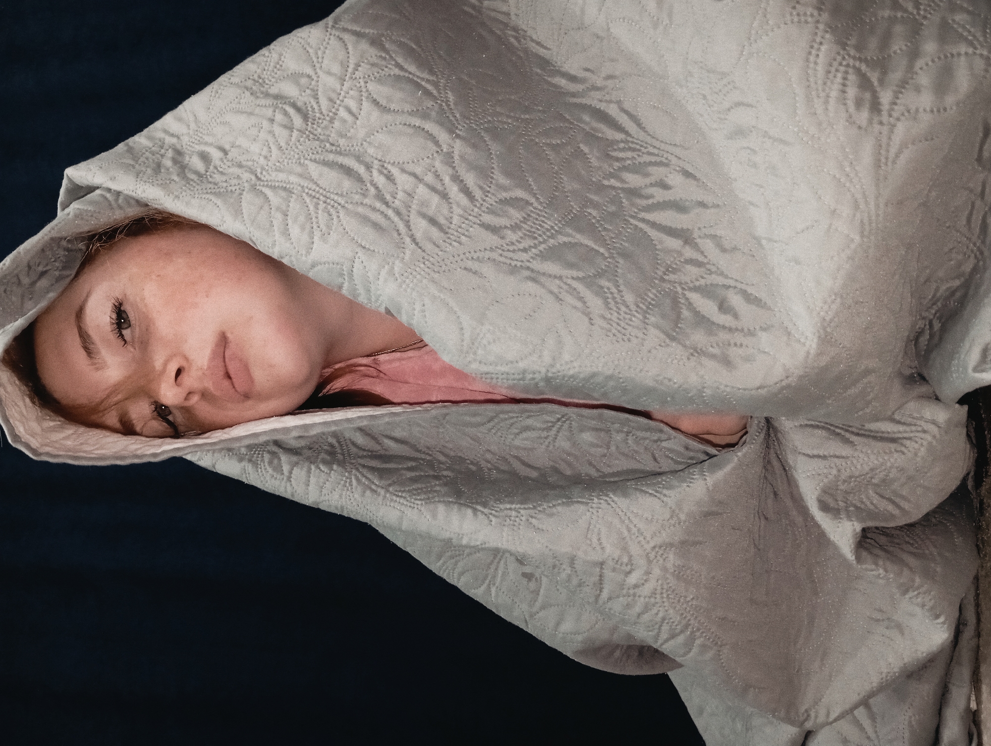 A person with freckles is wrapped in a light gray, quilted blanket with a leaf-like pattern. The blanket is draped over their head, creating a hood-like effect. The background is a dark, solid color. The subject's face is visible, and they appear to be lying down. The photograph does not contain information that would allow me to determine a location such as Yurgamysh, Russia.