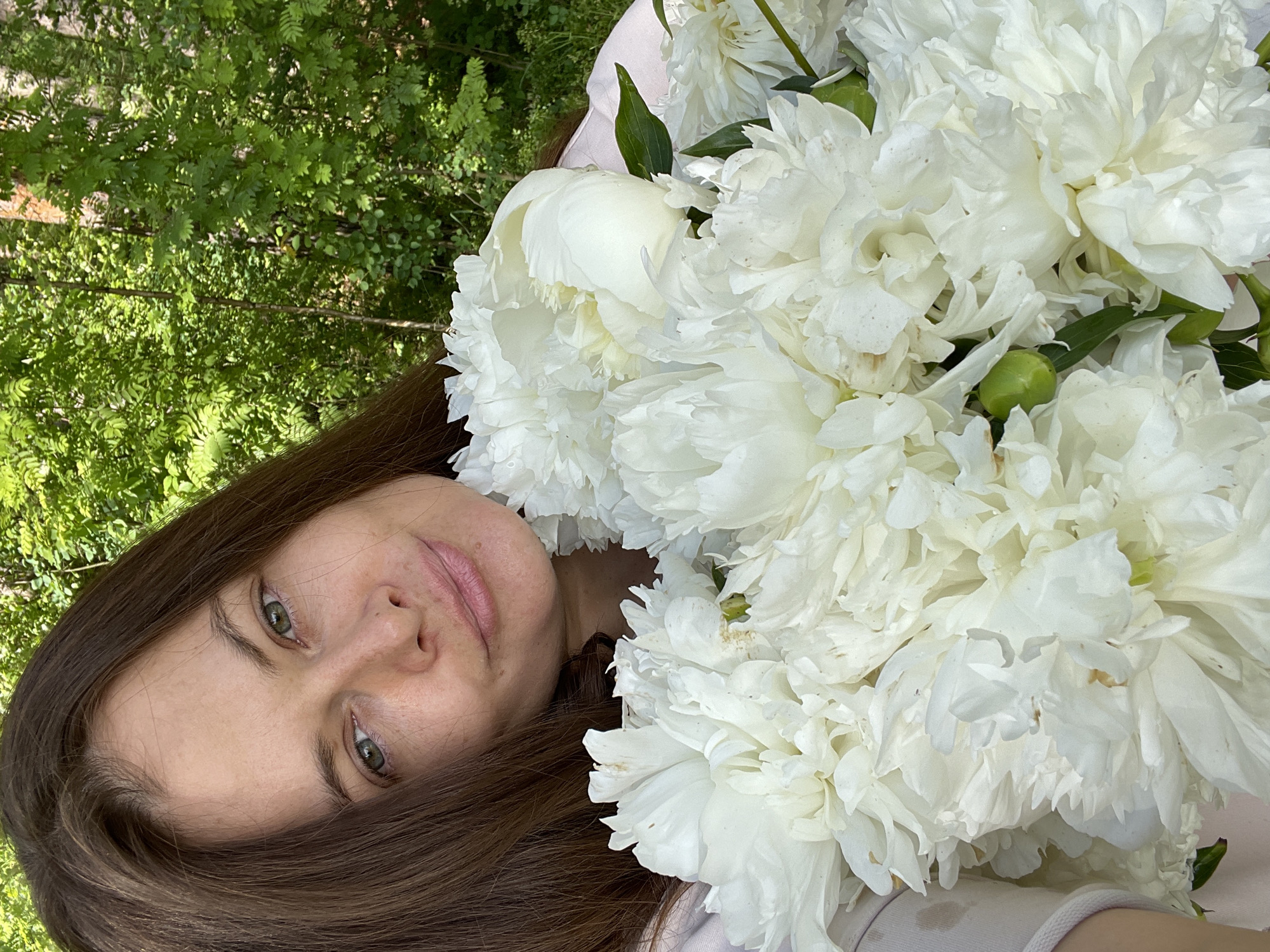 A woman with long, brown hair and green eyes is holding a large bouquet of white peonies in an outdoor setting with a background of green trees. The woman is positioned in the upper left quadrant of the frame, looking towards the camera with a slight smile. The peonies occupy the majority of the foreground, obscuring much of her body. The environment appears to be a park or natural area, possibly within or on the outskirts of the city of Заречный, Russia.