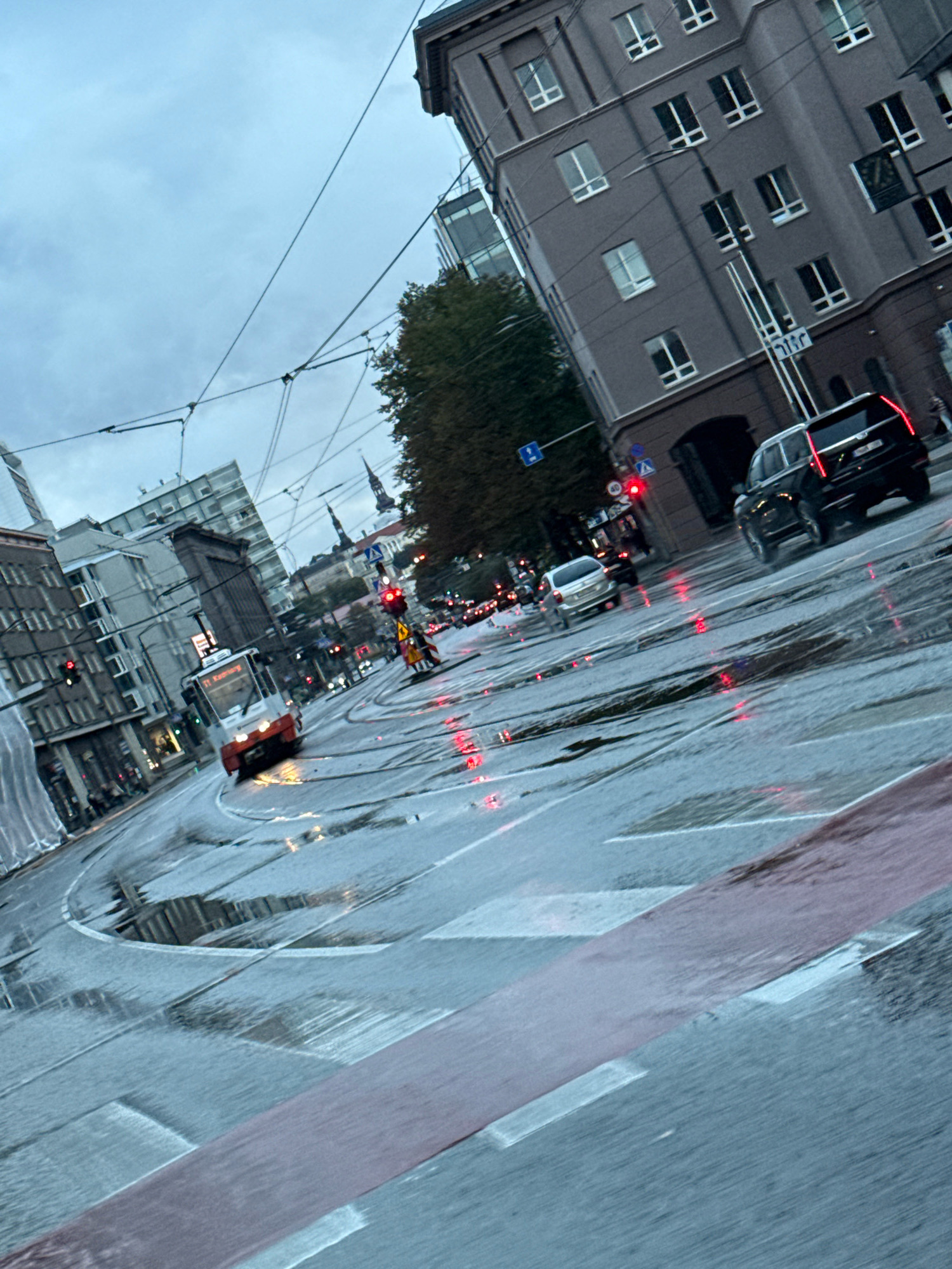 The image captures a street scene in Tallinn, Estonia, during a rainy day. A tram is visible on the wet tracks, illuminated by streetlights and vehicle headlights. Several cars are also present, navigating the slick road. Buildings line the street, some appearing to be older architecture, while others are modern structures. The sky is overcast, contributing to the dim lighting. Puddles of water on the road reflect the surrounding lights, creating a blurred and somewhat abstract effect. The overall impression is of a busy urban environment dealing with inclement weather.