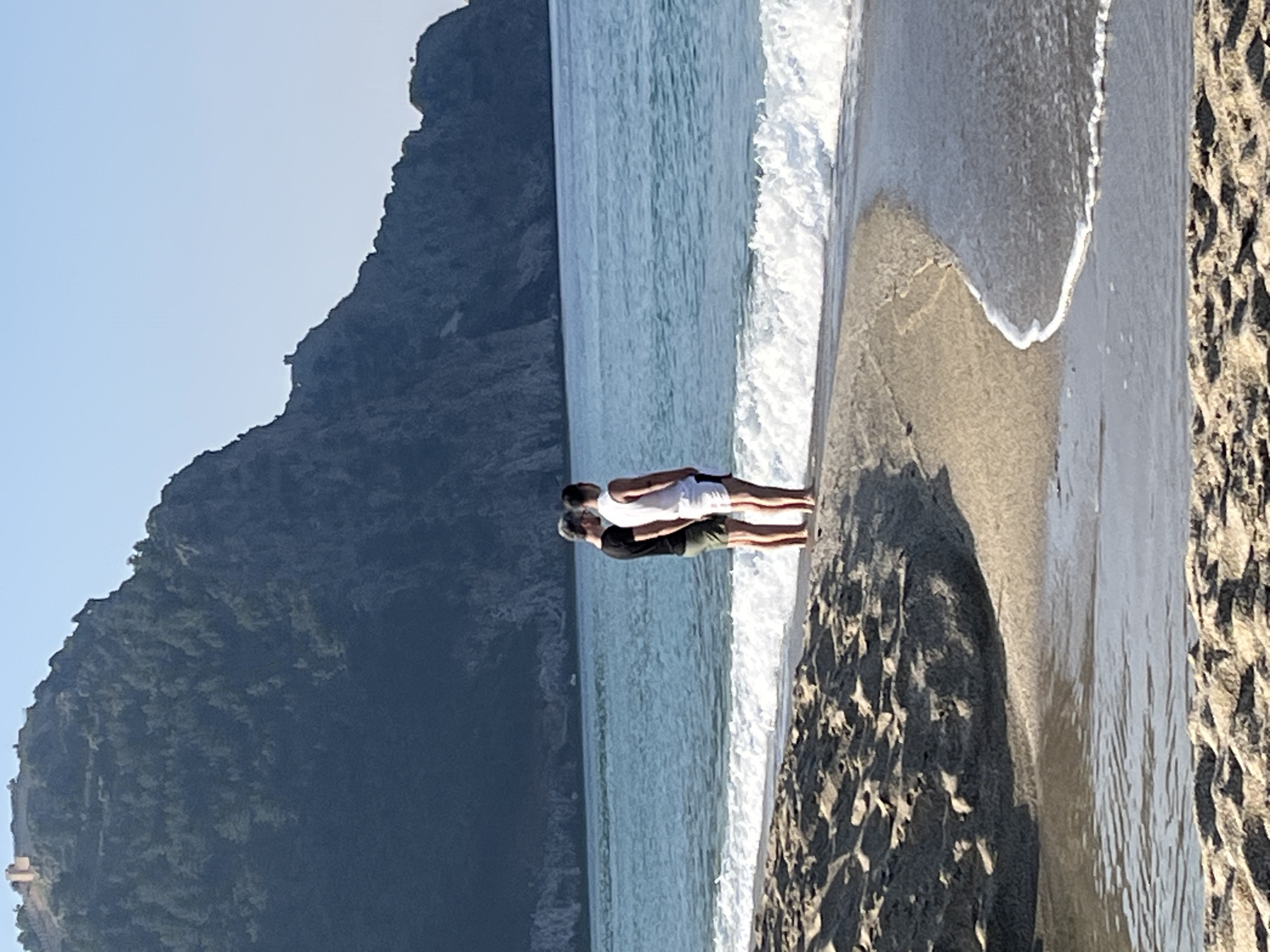 Here's a detailed description of the image:
The image shows a coastal scene, likely a beach, with a body of water (presumably the sea or ocean) and a large, rocky landmass or mountain in the background. The sky is light and clear.
In the foreground, there is a beach with sand. There are two people standing on the beach near the water's edge. One person is wearing a white top and the other is wearing a darker top. They appear to be standing close to each other.
The water is relatively calm with some visible waves or ripples. The background features a large, green, rocky hill or mountain that dominates the landscape.
Based on the location and features, this image may be taken near Alanya, Turkey, given that the landscape and coastal features align with that region. The large rock formation could be characteristic of the Alanya peninsula. The lighting indicates it is likely daytime.