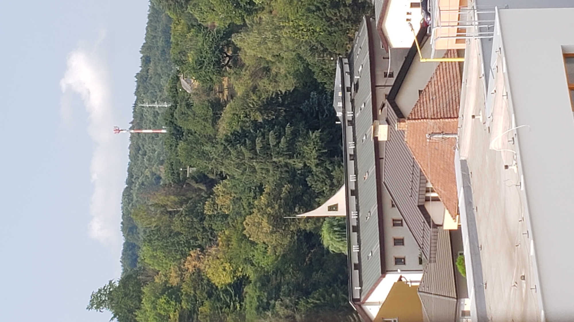 The image shows a view of a town or city with buildings and a wooded hill in the background. A red and white striped telecommunications tower is visible on the top of the hill. In the foreground, several buildings with different roof styles are present. One building has a distinctive white, sail-like structure on its roof, possibly part of a church or a modern architectural feature. The sky is partly cloudy. The overall scene appears to be a residential or mixed-use area with surrounding nature.