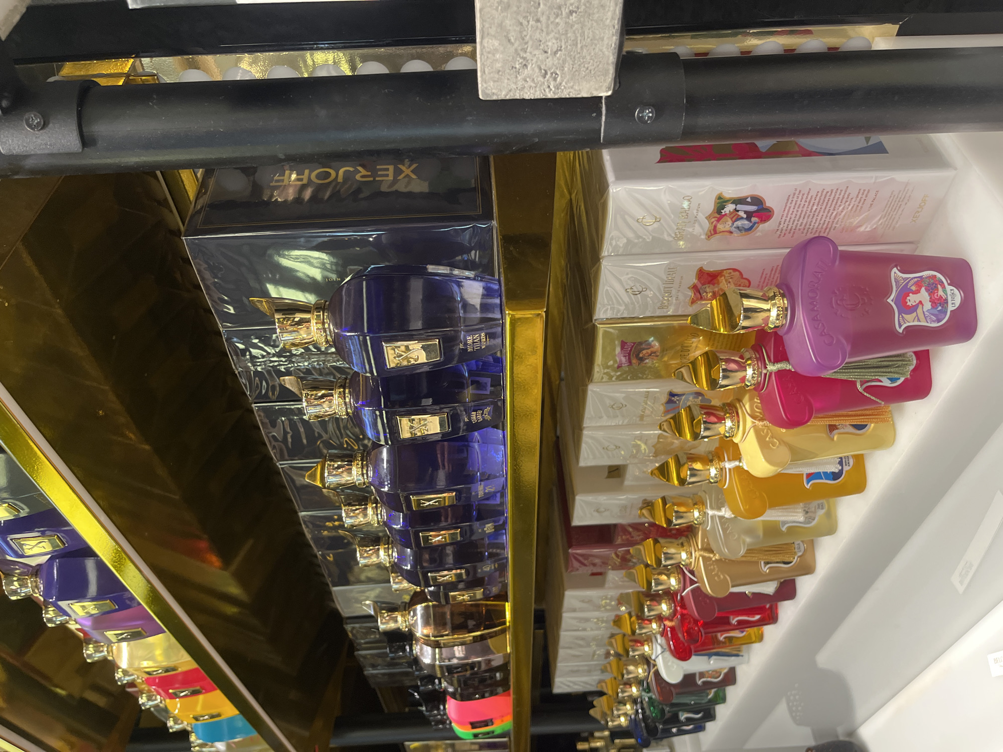 A shelf in what appears to be a retail environment displays numerous bottles of perfumes from the brands Xerjoff and Casamorati. The bottles are arranged neatly in rows and have various colors including blue, pink, red, yellow, and others. Some of the boxes and bottles bear text such as "More Than Words," "Re D'ulivo," "Bouquet Ideale," "Gran Ballo", and "Li Tosch". A black metal bar is visible at the top of the image. The location seems to be a store, possibly a perfume shop, located in Sofia, Bulgaria.
