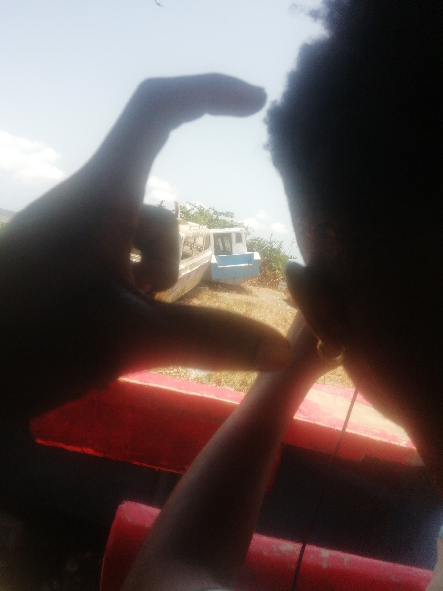 A blurry shot taken from a vehicle (likely a car) in Unknown, Unknown shows a person framing two abandoned boats on land with their hands. The person's face is only partially visible, obscured by dark hair and a hand. The left hand is more clearly visible, silhouetted against a bright sky, creating a rough heart shape with the other hand. Through the frame, two beached boats of different sizes and colors (white and light blue) are visible on a patch of grass. The foreground includes the vehicle's red interior trim. The scene is captured in broad daylight.