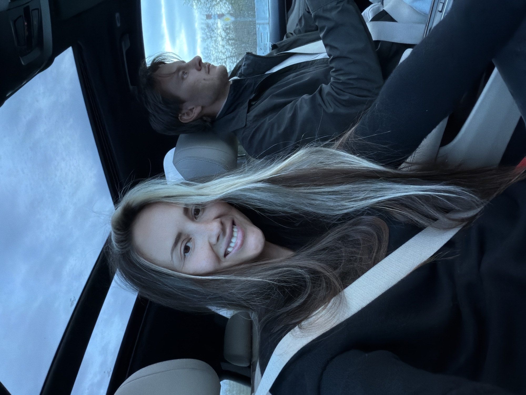 This image captures two individuals inside a car, likely in Moscow, Russia, on an overcast day. The photograph is taken from a low angle, looking upwards through a panoramic sunroof that reveals a cloudy sky.
In the foreground, a woman with long, multi-toned hair (featuring blonde highlights and darker roots) is smiling broadly at the camera, appearing to take a selfie. She is wearing dark clothing and has a light-colored seatbelt draped across her chest, securing her in the seat. Her expression is cheerful and engaging.
Behind her, a man with short, dark hair is visible, looking upwards and away from the camera. He is wearing a dark jacket over a lighter shirt and also has a light-colored seatbelt across him. His expression appears neutral or contemplative.
The car's interior features light-colored seats and headrests, contrasting with the dark roof lining. The soft, diffused lighting suggests it is daytime but without direct sunlight. Through a side window in the background, some blurry shapes, possibly trees or buildings, are indistinctly visible, indicating the car is in motion or parked in an outdoor setting. There is no visible text within the image.