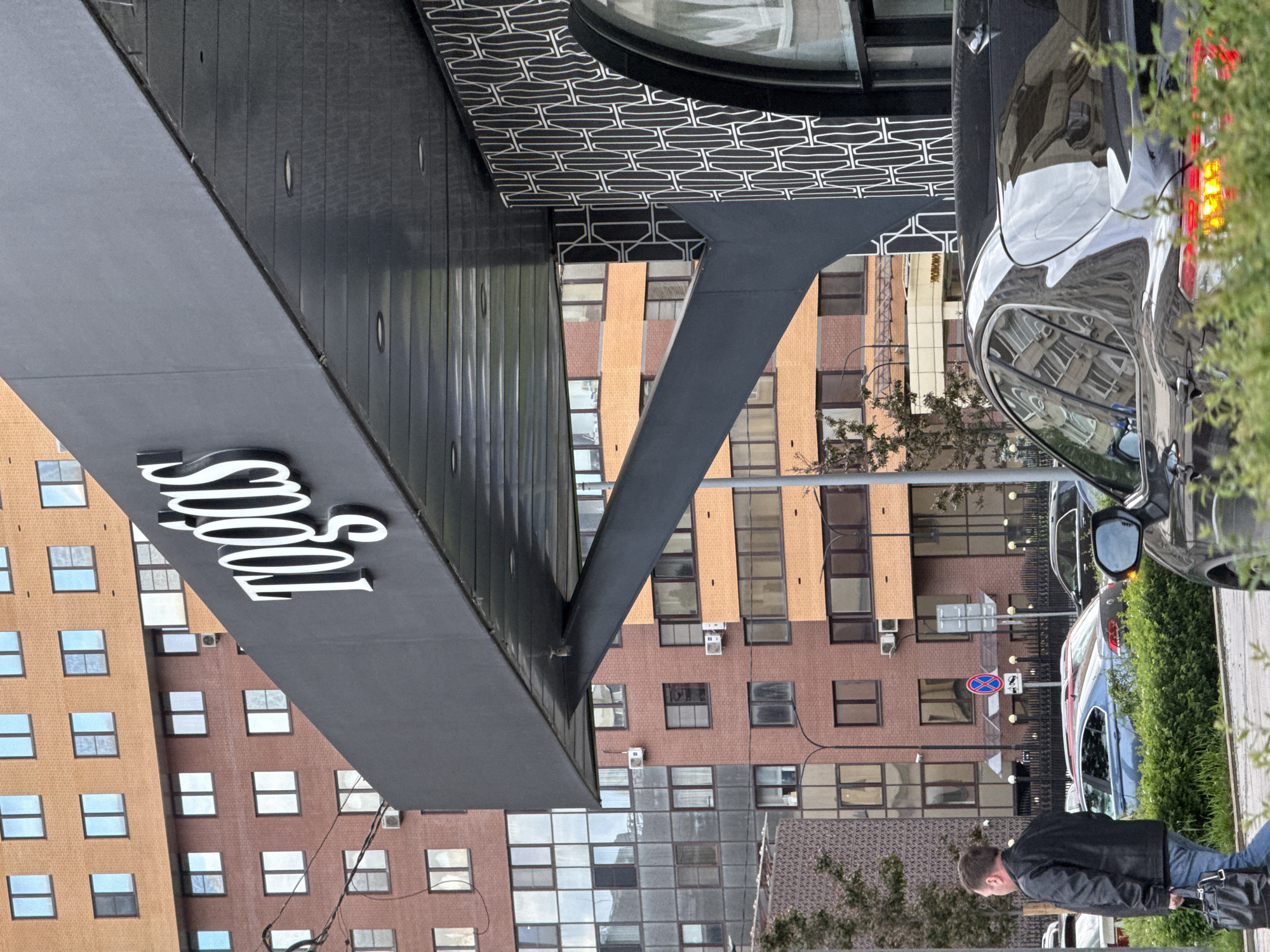 The image captures a street scene in Moscow, Russia. Buildings with repetitive window patterns dominate the backdrop, featuring a mix of orange-brick and glass facades. A black cantilevered awning bearing the word "Todos" in a stylized white font juts out from one of the buildings. Supporting the awning is a dark gray, angled support beam. A patterned ceiling feature is visible above.
Parked cars, including a dark-colored SUV, line the street, partially obscured by greenery. A man wearing a black jacket and blue jeans is walking along the sidewalk. A street sign is visible in the background with white, dark blue, and red markings. The composition is captured at a rotated angle, and the overall lighting suggests an overcast day.