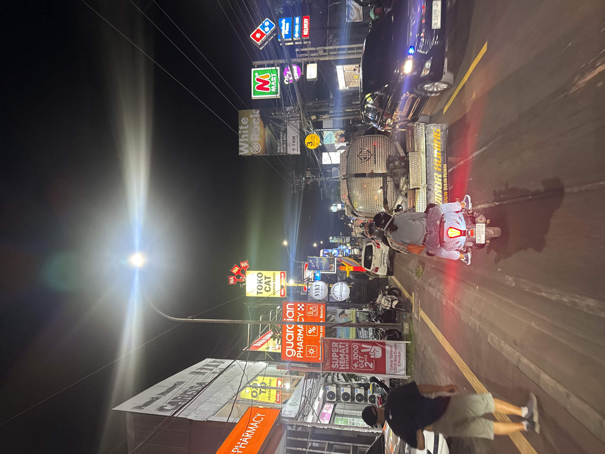 At night in Kuta Selatan, Indonesia, the image shows a street scene with multiple businesses, including a Carissa Pharmacy, Guardian Pharmacy, Toko Cat Warna Abadi paint store, and a White Rock beachfront club. Signs for M Mart, NYKY Beauty Salon, Domino's Pizza, and other commercial establishments are visible. A street lamp is illuminating the scene with lens flare. A person is seen walking on the sidewalk. A motorcycle with illuminated taillights is on the road next to a truck. A yellow line marks the lane on the street. Other motorcycles are parked along the sidewalk. The night sky is visible, with numerous electrical wires crisscrossing above the street.