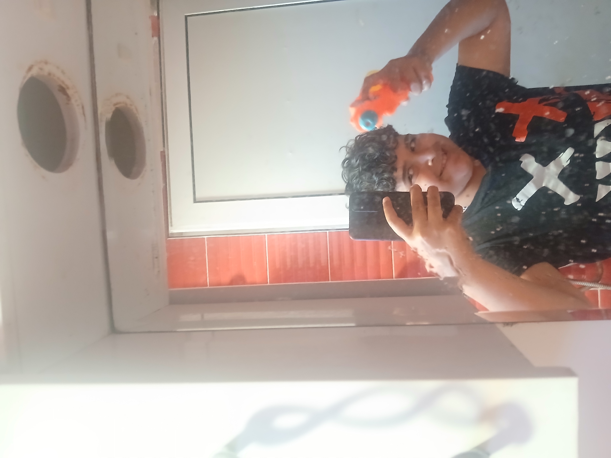 A young person with curly dark hair is taking a selfie in a bathroom. The person is holding a black smartphone in their left hand. In their right hand, they hold an orange and blue toy water gun. The person is smiling. The person is wearing a black t-shirt with white and red X designs. Behind the person is a mirror reflecting the image. Above the mirror is a white cabinet with two circular openings. Red tiles are visible along the wall. The bathroom is located in Ignatievo, Bulgaria.