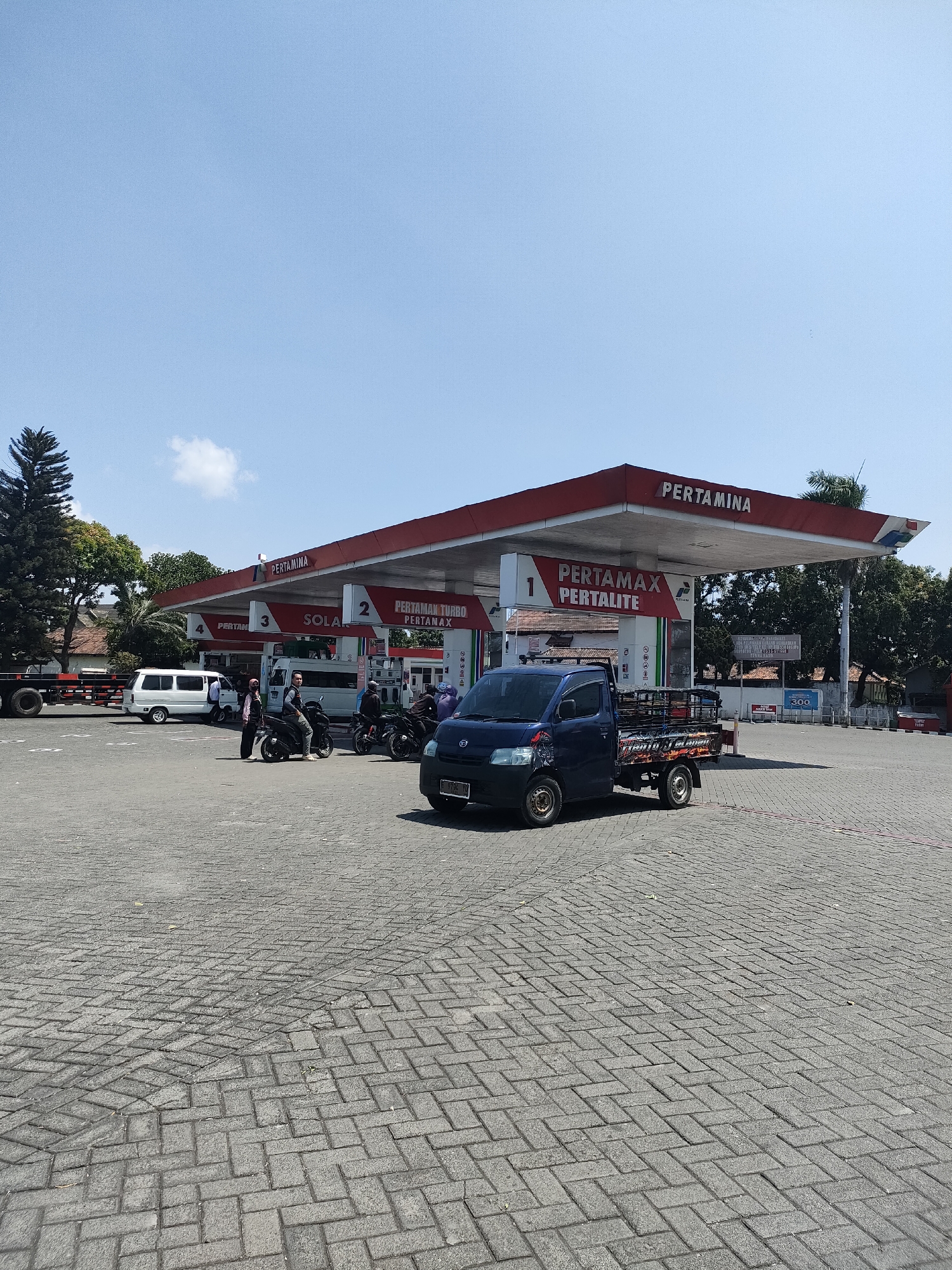 The image depicts a Pertamina gas station in Curahdringu, Indonesia. The gas station features a red and white canopy covering multiple fuel pumps, with signage indicating "PERTAMINA," "PERTAMAX," "PERTALITE," and "SOLAR." Several vehicles are present, including a blue pickup truck with a cage in the back, a white van, and multiple motorcycles. People are seen near the motorcycles, some appearing to be refueling or interacting. The ground is paved with interlocking bricks. Trees and some buildings are visible in the background under a clear blue sky.