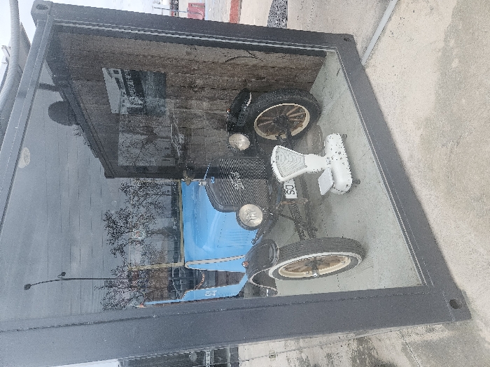 A light blue vintage automobile, identifiable as a Ford Model T, is displayed within a large, rectangular, dark-framed glass exhibit case situated on a paved concrete surface in the city of Tudela, Spain. The vehicle features a light blue body, black fenders, dual circular headlights, and spoked wooden wheels with white-sided tires. A license plate partially visible on the front reads "SO." Inside the case, positioned on the ground in front of the car, is an antique white mechanical scale equipped with a circular dial and a flat weighing platform. A dark, textured panel serving as a backdrop to the vehicle displays an affixed black and white historical photograph. Reflections on the glass of the exhibit case show an overcast sky, a street lamp, and bare deciduous trees. In the background, beyond the display's glass and frame, red and white traffic barriers and a large white tanker truck are partially visible, indicating an outdoor, urban or roadside environment. No individuals are present in the scene.