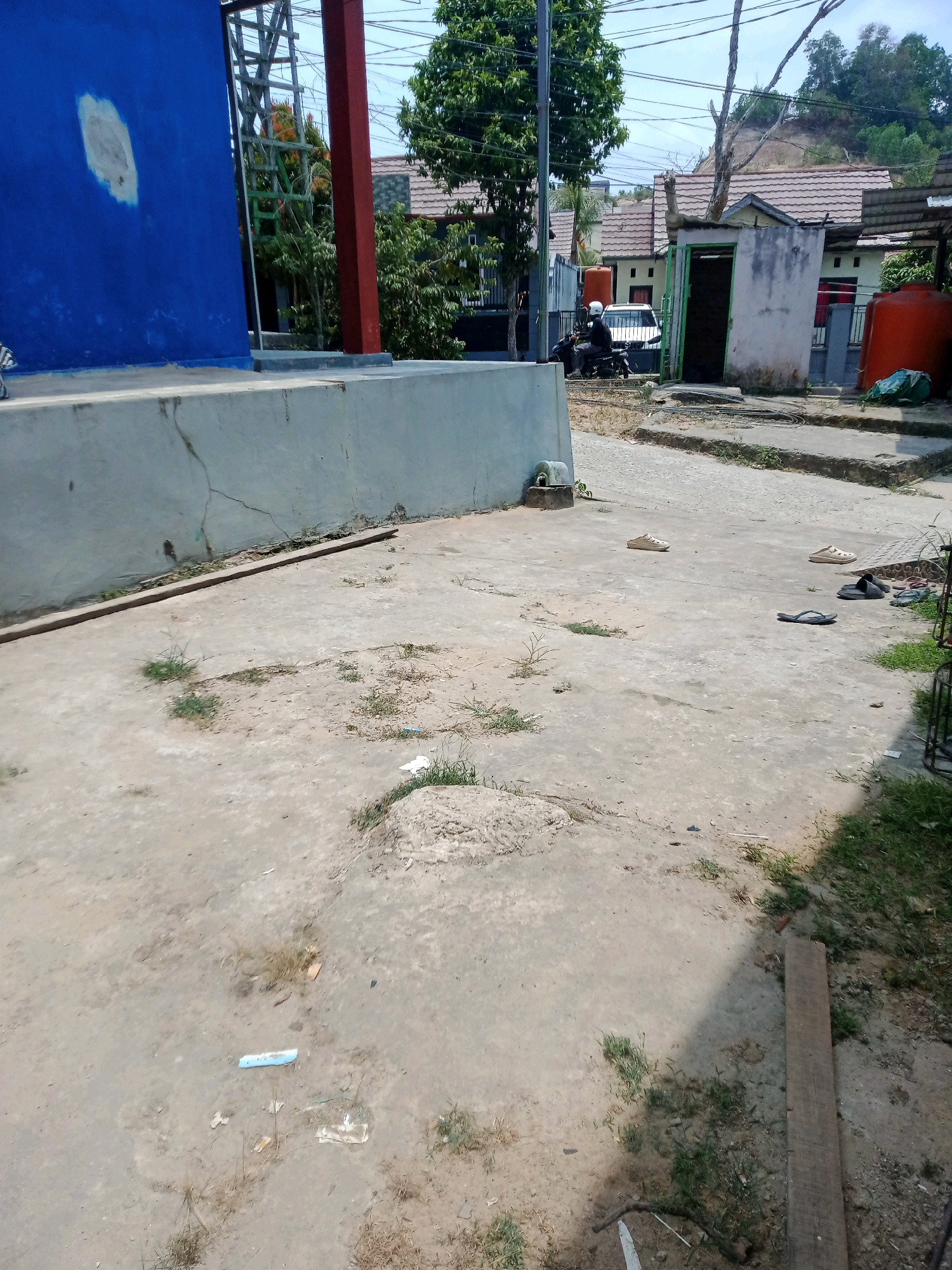 The image depicts an outdoor scene in what appears to be a residential area in Samarinda, Indonesia. In the foreground, a concrete pavement is visible, with patches of dry grass and some debris scattered across it. Further back, a low concrete wall runs horizontally across the frame. To the left, a portion of a building with a bright blue wall and a faded circular patch is seen. To the right of the blue building, there is a metal scaffolding structure.
Beyond the concrete wall, a street or pathway leads into a neighborhood. Several houses with tiled roofs are visible, suggesting a suburban or densely populated residential zone. In the middle ground, a person is riding a motorcycle, wearing a white helmet. A white car is parked behind the motorcycle. Adjacent to the houses, there is a small, open-fronted structure with a weathered exterior and a green door frame. A large orange water tank is also present on the right side of the image. Trees and power lines are visible in the background, and a hillside covered in vegetation is in the distance. The overall impression is of a fairly typical urban or peri-urban environment.