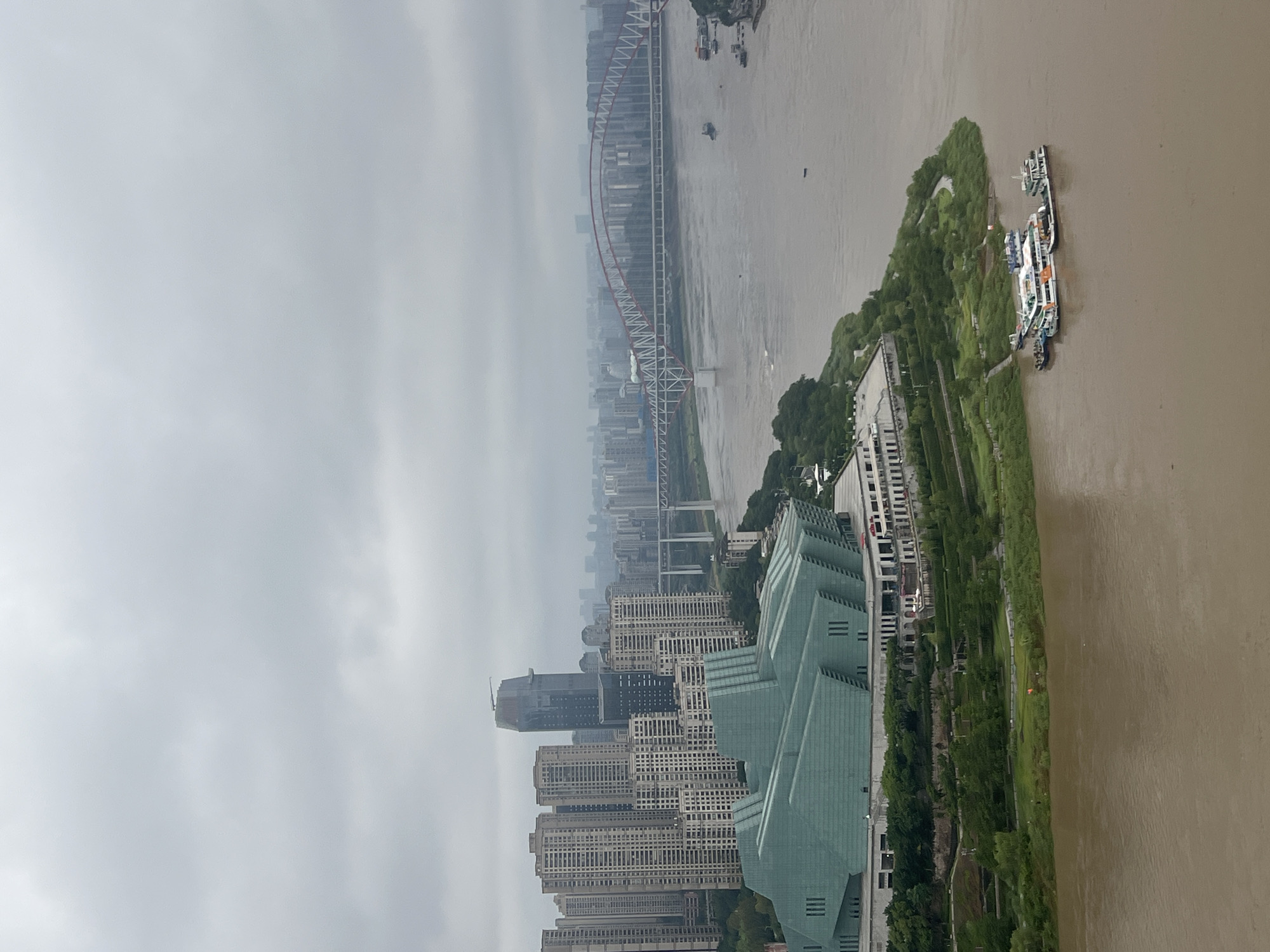 This is an aerial, high-angle, full shot of a river in Yuzhong District, China. The river is wide and murky brown, with several boats docked along its edge. In the foreground, a cluster of boats is moored together. To the left, a large, modern, green-roofed building with a distinctive angular design sits on the riverbank, surrounded by lush greenery. Beyond this building, a densely populated urban landscape unfolds, with numerous tall, modern skyscrapers. A long, arched bridge with red and white supports spans the river in the middle ground, connecting the two sides of the city. The sky is overcast and gray, indicating a cloudy day.