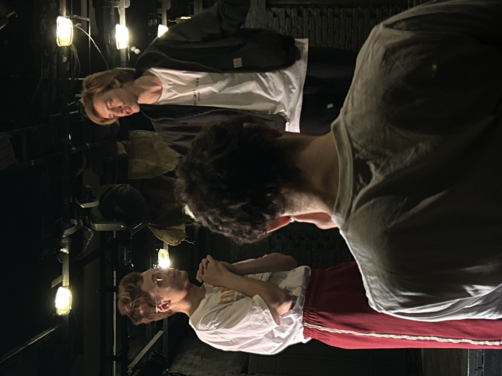 In an indoor setting that appears to be a stage or rehearsal space, three young men are visible. The man on the left, wearing glasses, a white t-shirt, and red pants with a white stripe, stands with his hands clasped. To his right, a man with blond hair, wearing a white t-shirt and a dark jacket, is speaking. In the foreground, a third man, seen from behind, is wearing a light-colored t-shirt. The background features stage lighting and what appears to be theatrical set pieces, possibly made of burlap or similar material, suspended from the ceiling. The overall lighting is dim, with spotlights illuminating the individuals.