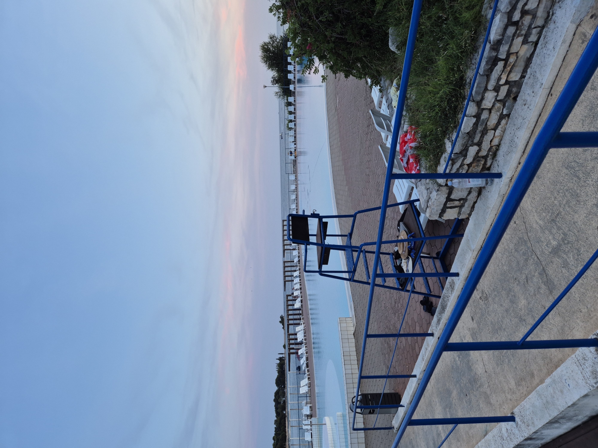 The image depicts an outdoor swimming pool area at dusk in Katoro, Croatia. The large, rectangular pool is filled with calm, light blue water and is surrounded by white lounge chairs. A blue lifeguard chair with steps stands prominently in the foreground, positioned to overlook the pool. The deck surrounding the pool is paved with reddish-brown bricks. A blue railing with white concrete supports runs along a pathway in front of the pool area. Beyond the pool, a body of water, likely the Adriatic Sea, is visible, with a distant shoreline dotted with trees and structures. The sky exhibits a soft gradient of blues and pinks, indicative of twilight. A small cluster of green bushes and some scattered white lounge chairs are visible on the right side of the frame, near a stone wall. The overall scene suggests a peaceful, recreational setting at the end of the day. There are no people visible in the image.