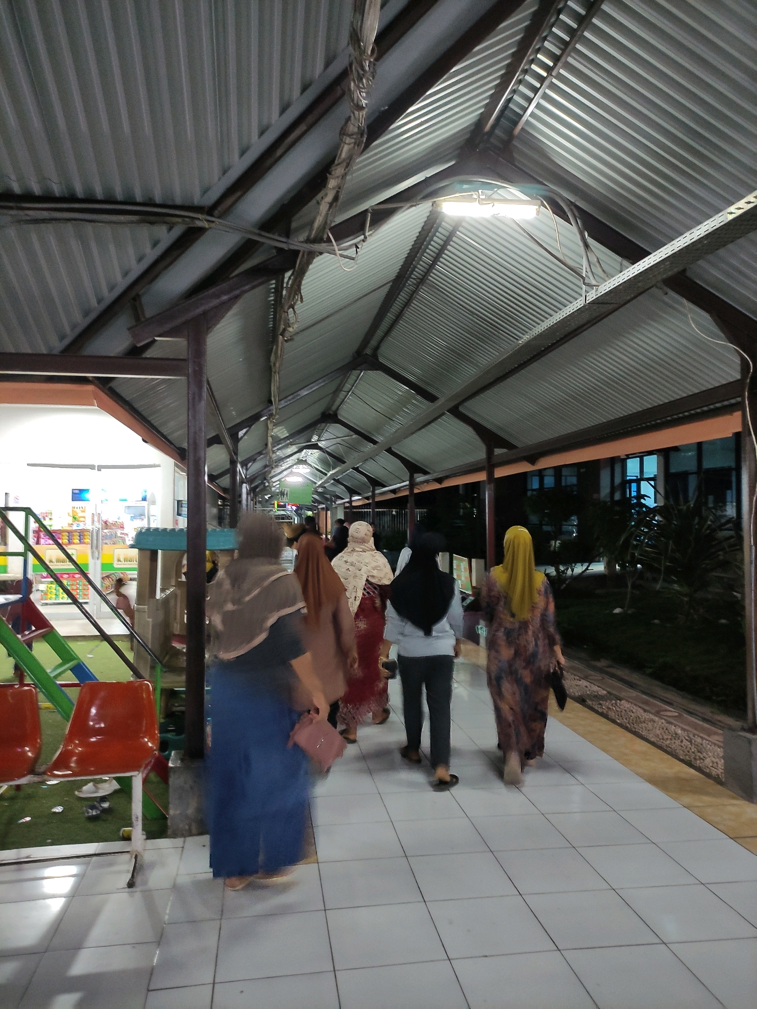 The image depicts an outdoor walkway covered by a corrugated metal roof supported by dark brown metal posts and beams. The walkway is paved with white square tiles and appears to be at dusk or night, with overhead fluorescent lights illuminating the scene. Several people, mostly women wearing hijabs and long dresses or skirts, are walking away from the camera down the covered path. On the left side of the walkway, there is an open-sided building with shelves displaying goods, suggesting it could be a shop or convenience store. Adjacent to this building is a children's play area with a red slide and a blue play structure, and two bright orange chairs. The walkway extends into the distance, with greenery and buildings visible on the right side, some with blue-tinted windows. The context provided suggests this scene is located in Bulukumba, Indonesia.