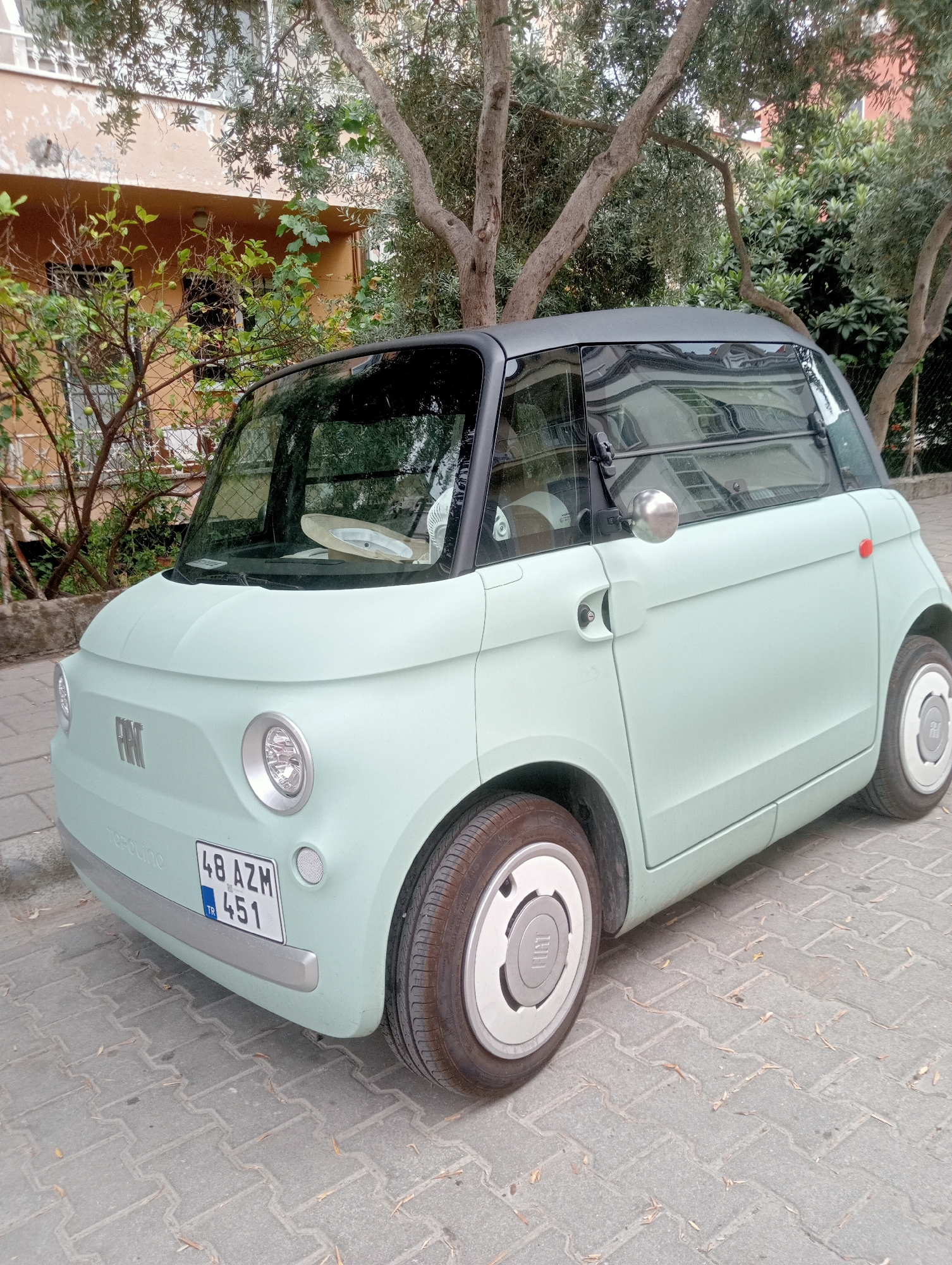 A light green, retro-styled Fiat Topolino electric car is parked on a paved sidewalk in Fethiye, Turkey. The car has a black roof and is facing slightly to the left. A Turkish license plate, reading "48 AZM 451", is visible on the front bumper. The background shows a building with an orange facade and trees with dense foliage. The ground is covered in interlocking gray paving stones.