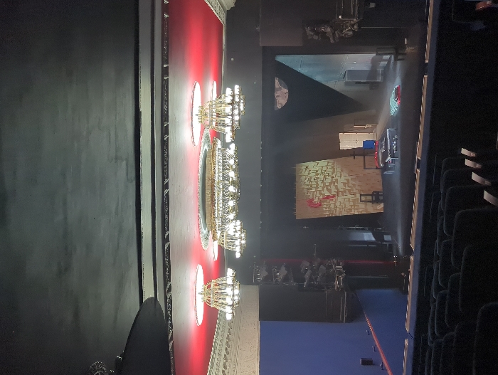 The image depicts the interior of a theater in Moscow, Russia, viewed from the audience seating towards the stage. The auditorium ceiling is dark, below which a red-lit section features three ornate, gold-toned chandeliers with multiple bright, white-light-emitting bulbs. A larger central chandelier is flanked by two smaller ones. Rows of dark-colored, upholstered theater seats, appearing empty, occupy the foreground.
The stage is dimly lit with spotlights illuminating specific areas. On the left side of the stage, a tall, solid blue backdrop is visible, across which a diagonal red line or beam extends. A vertical truss supporting multiple stage lighting fixtures stands nearby. Center-stage features a large, light-colored panel, possibly plywood, displaying a textured, block-like pattern. Overlaid on this panel is an abstract, red, stylized design. In front of the panel, two dark, simple chairs are positioned. A low, rectangular platform, covered in dark fabric, holds several white items, possibly textiles. To the right of the platform, a pile of colorful fabrics, predominantly teal and red, rests on the stage floor. The right side of the stage reveals multiple dark doorways or entrances and an upper semi-circular opening on a dark wall, with a dark structural element in the foreground. No individuals are present within the scene.