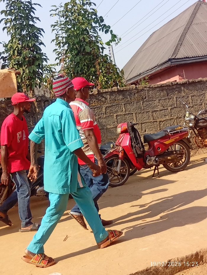 Three men walk down a street alongside parked motorcycles. The first man, wearing a red cap and shirt with jeans, walks to the left. The second man is wearing a colorful hat and a turquoise outfit. The third man is wearing a red cap and red and white striped shirt. A low brick wall is in the background with a building behind it. Trees are also in the background. The date "19/12/2025" is at the bottom of the image.