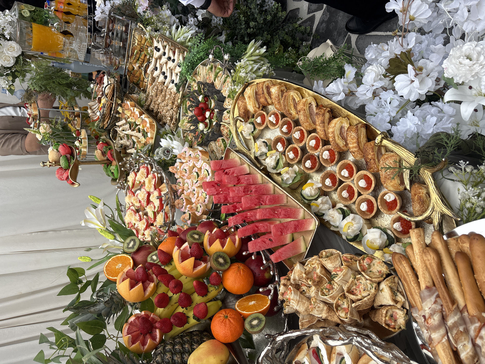 A high-angle, close-up shot reveals a lavish catering display. The setup includes an assortment of fruit like watermelon, oranges, grapefruits, strawberries, bananas, kiwi, pineapple, and mango. There are also various small appetizer items such as mini tarts with red caviar, puff pastries, and wraps. The setting appears to be a reception or party given the large array of food. The location cannot be determined from the visual data, nor that it is in Yerevan, Armenia. There is no other information available about the presence of people.