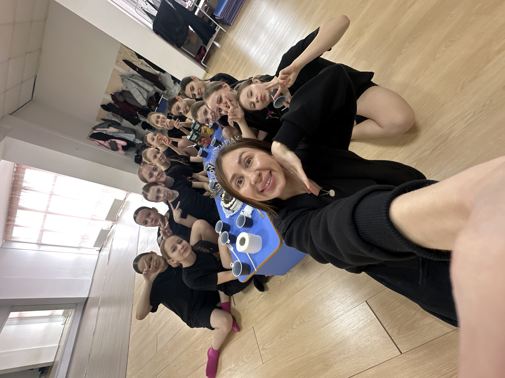 A selfie captures a group of approximately 13 people, mainly young females, indoors in a location identified as Votkinsk, Russia. All are dressed in dark clothing, and some are making peace signs with their fingers. A small, blue table is positioned in front of them, containing toy cups, a roll of toilet paper, and a boxed item. The background includes a wall-mounted rack filled with clothing, and the room appears to have wooden floors and light-colored walls.