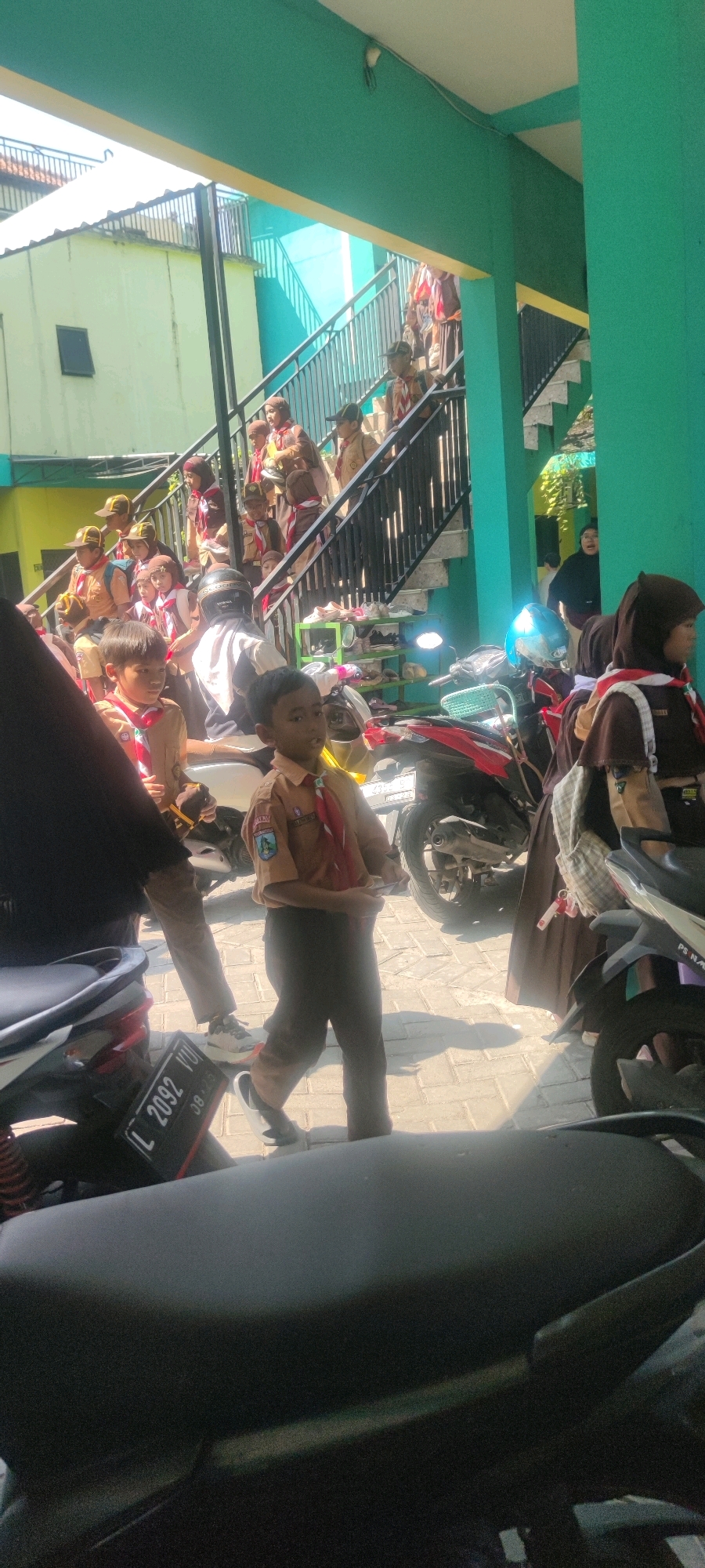 In the city of Banjarsari, Indonesia, a group of schoolchildren in scout uniforms are gathered outdoors, near a building with a green facade and yellow trim. A set of outdoor stairs, lined with a black railing, dominates the midground, with many students ascending or descending. Some students are seen standing or sitting on the ground level, near parked motorcycles. In the foreground, a motorcycle with a license plate reading "L 2092 VU" is partially visible. The children are mostly wearing tan scout shirts with red neckerchiefs, and some are wearing hats. The overall scene suggests an outdoor school activity or assembly.