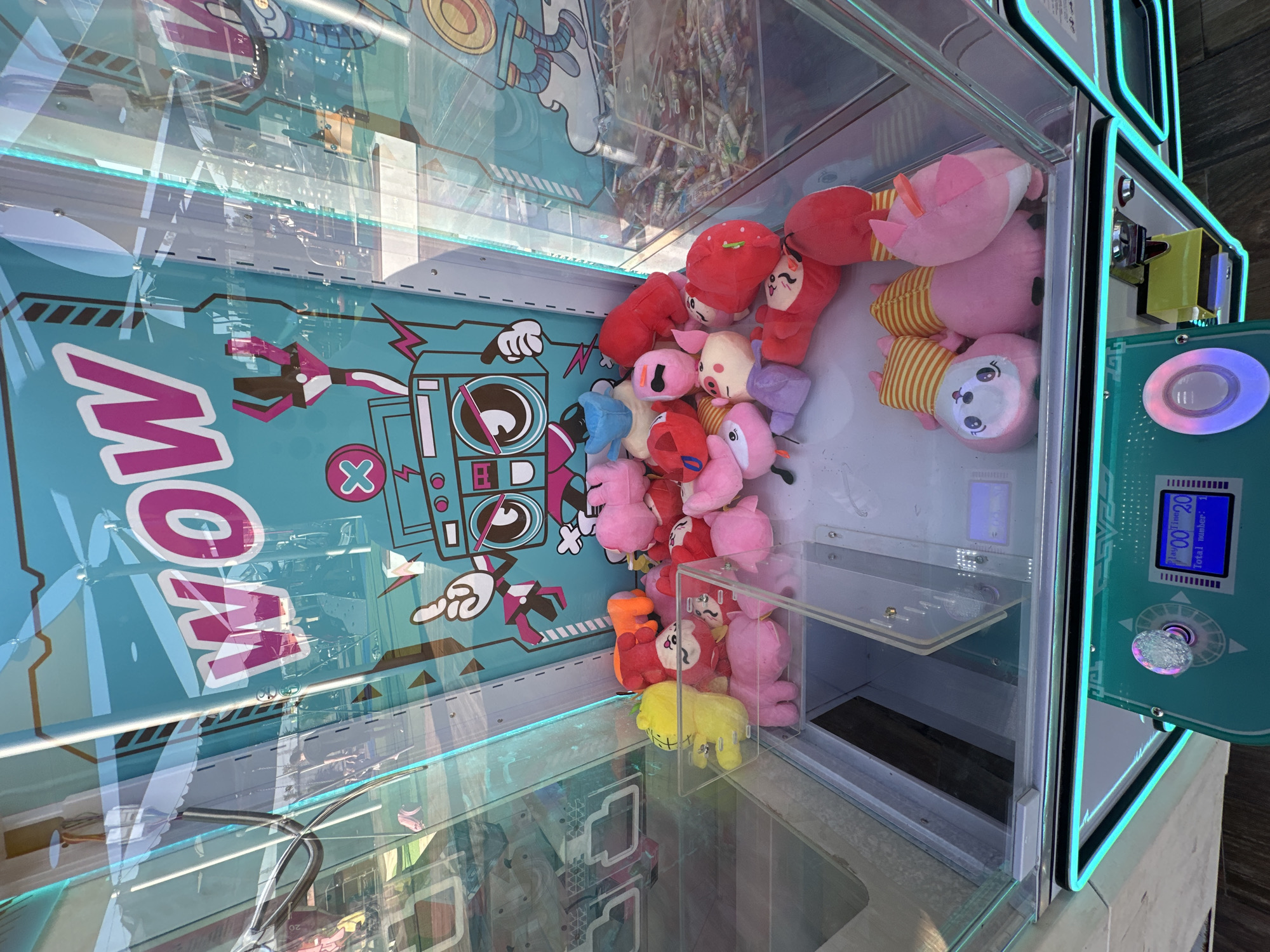 This image depicts the interior of a colorful claw machine arcade game filled with plush toys. The background features a vibrant teal graphic with the word "WOW" in large pink letters and cartoonish illustrations of a boombox and a robot. Various plush toys, predominantly pink, red, and yellow, are piled inside the machine, some are characters with faces and striped hats. In the foreground, a control panel with a digital display shows "Time: 20" and "Total number: 1," and a translucent joystick. The overall environment suggests a brightly lit amusement or entertainment venue. The location provided is Hod HaSharon, Israel.