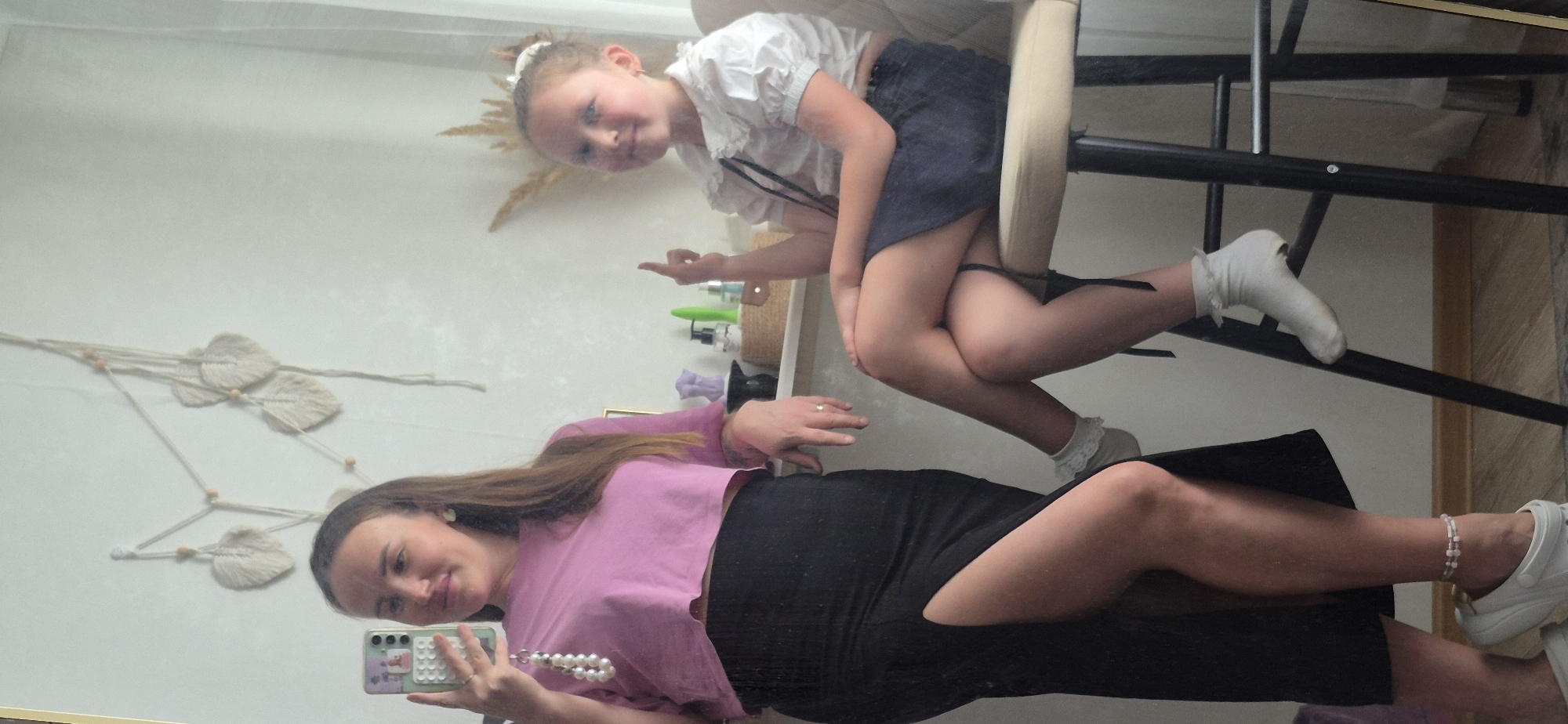 A reflection in a mirror shows a woman taking a selfie with a child sitting on a chair. The woman is wearing a pink crop top and a black skirt with a side slit, and she is holding a phone with a beaded strap. The child is wearing a white blouse and a navy skirt, and she is sitting on a beige chair with black legs. The background includes a white wall with macrame decorations and a shelf with various items.