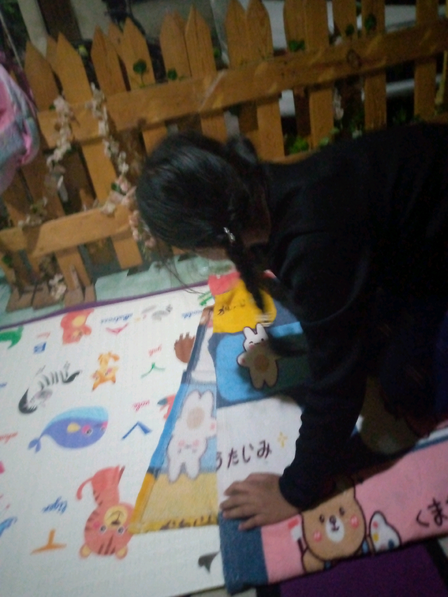 A young girl wearing a black top is in an indoor setting, possibly a bedroom or playroom. She is crouching over a colorful mat with cartoon animal designs, including a tiger, a whale, and several rabbits. She appears to be reaching for something on the mat. A wooden fence is visible in the background, decorated with small white flowers. The lighting is dim, suggesting it might be evening or the room is not well-lit. The location is identified as Sumberwono, Indonesia.