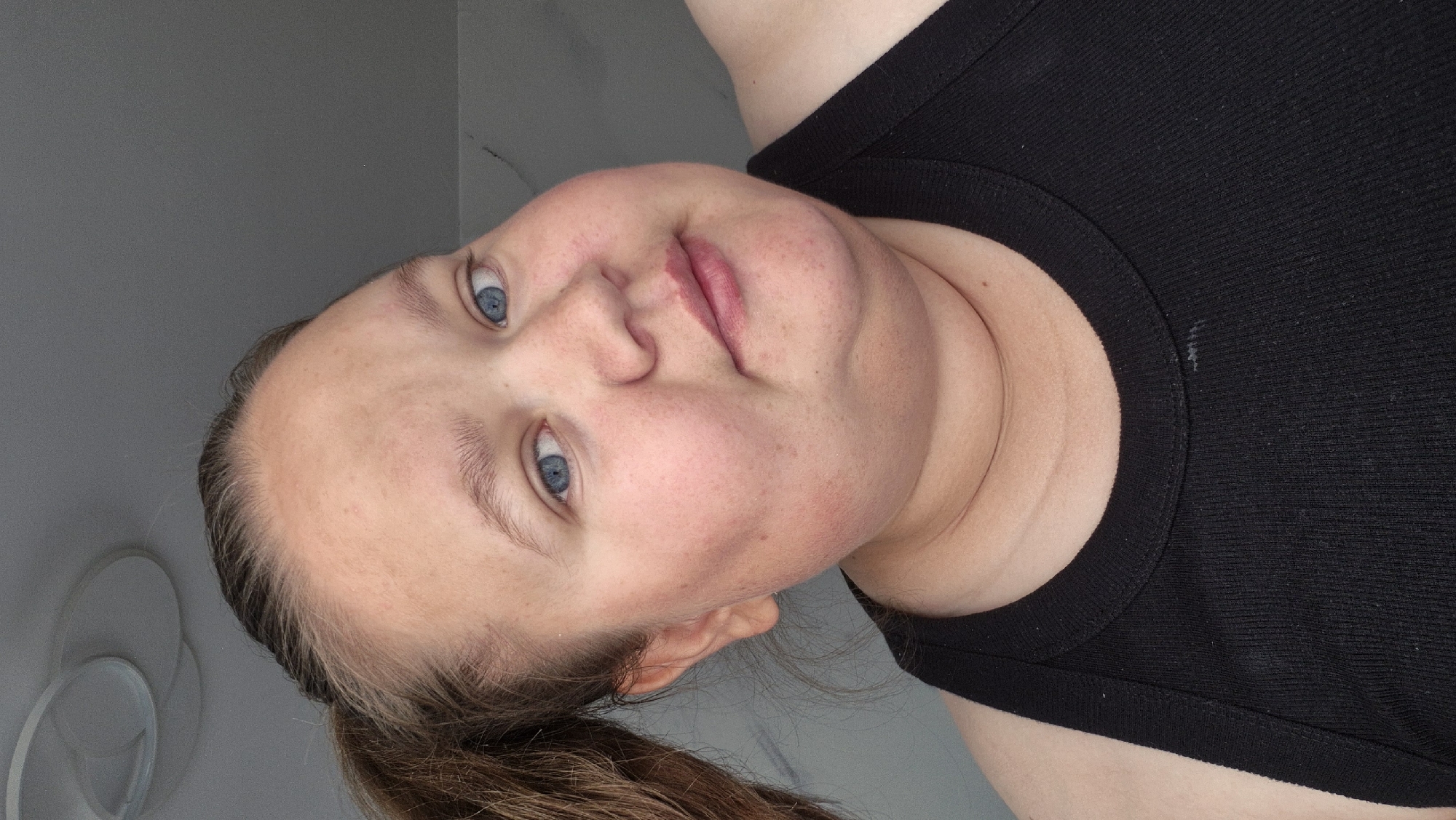 The image presents a close-up portrait of a female individual, captured from the upper chest upward. She has fair skin, with visible freckling across her forehead, cheeks, and chin, and prominent blue eyes that gaze directly forward. Her light to medium brown hair is pulled back into a ponytail, partially visible on the left side of the frame, with some lighter, possibly graying, strands near the hairline. Her face is round, and she displays a slight, closed-mouth smile. Subtle redness or blush is present on her cheeks, and folds are visible under her chin and along her neck. She is dressed in a black, ribbed tank top with a high round neckline. A small, light-colored smudge is discernible on the fabric of her top below the neckline. The background consists of a plain, light gray wall. In the upper left corner, a portion of a modern, multi-ringed ceiling light fixture is visible, indicating an indoor setting. The specific location, including the city of Chaikovsky, Russia, is not discernible from the visual information provided.