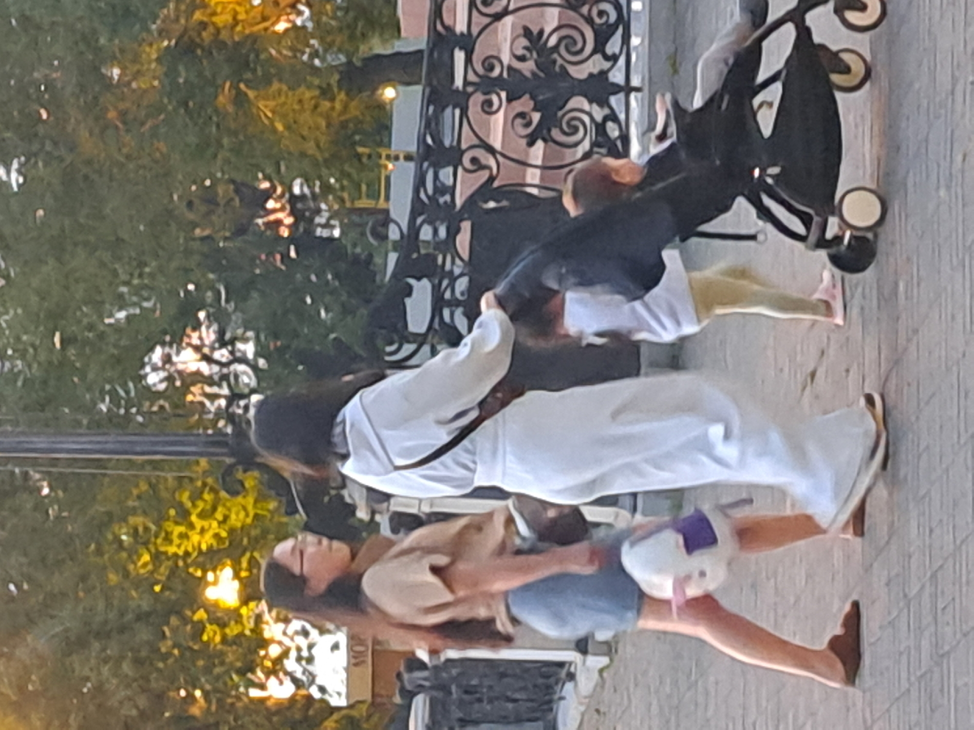 In Irkutsk, Russia, two women and two children are depicted walking on a paved pathway. The woman on the left is wearing a tan t-shirt and a denim skirt, carrying a stuffed rabbit with purple accents. The woman on the right is dressed in a white tracksuit. Behind them, a stroller containing a child is visible, with another child walking nearby. The background features trees with yellow and green foliage, suggesting it is autumn, and a decorative metal fence. The lighting indicates it is daytime.