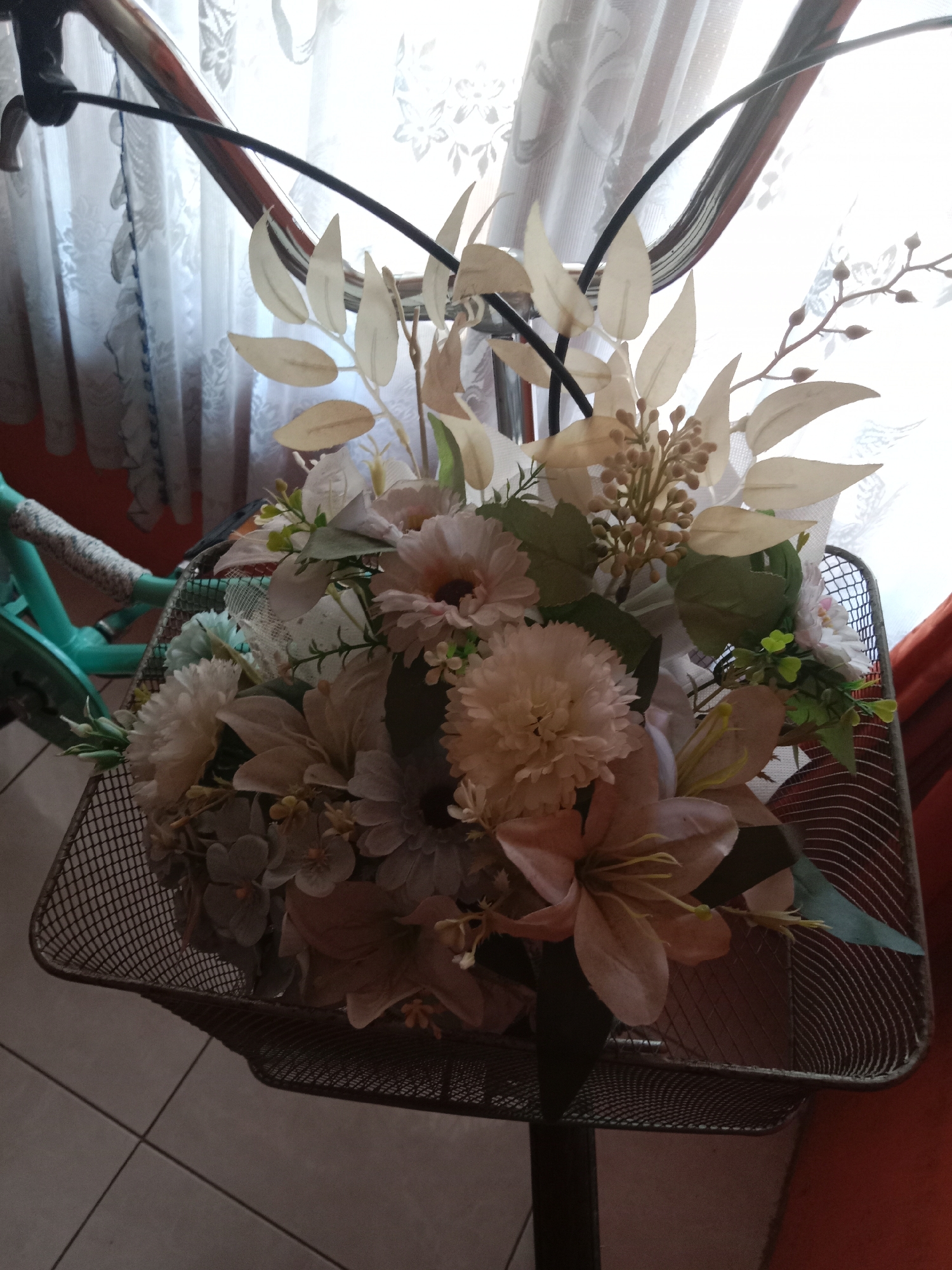 The image shows a close-up view of a bouquet of artificial flowers in a bicycle basket. The basket is made of dark metal mesh and is attached to the handlebars of a bicycle, which is partially visible on the left side of the frame. The bicycle frame is teal colored. The bouquet consists of various flowers in shades of cream, peach, and pale pink, along with some greenery and decorative branches with small buds. The background features a sheer white curtain with a floral pattern, and a glimpse of an orange curtain to the right. The lighting suggests it is daytime, with natural light filtering through the curtains. The floor appears to be tiled. The image is taken indoors, likely in a home environment. There are no people visible in the image. The scene does not provide any specific information that can link it to Kutosari, Indonesia, or any particular city.